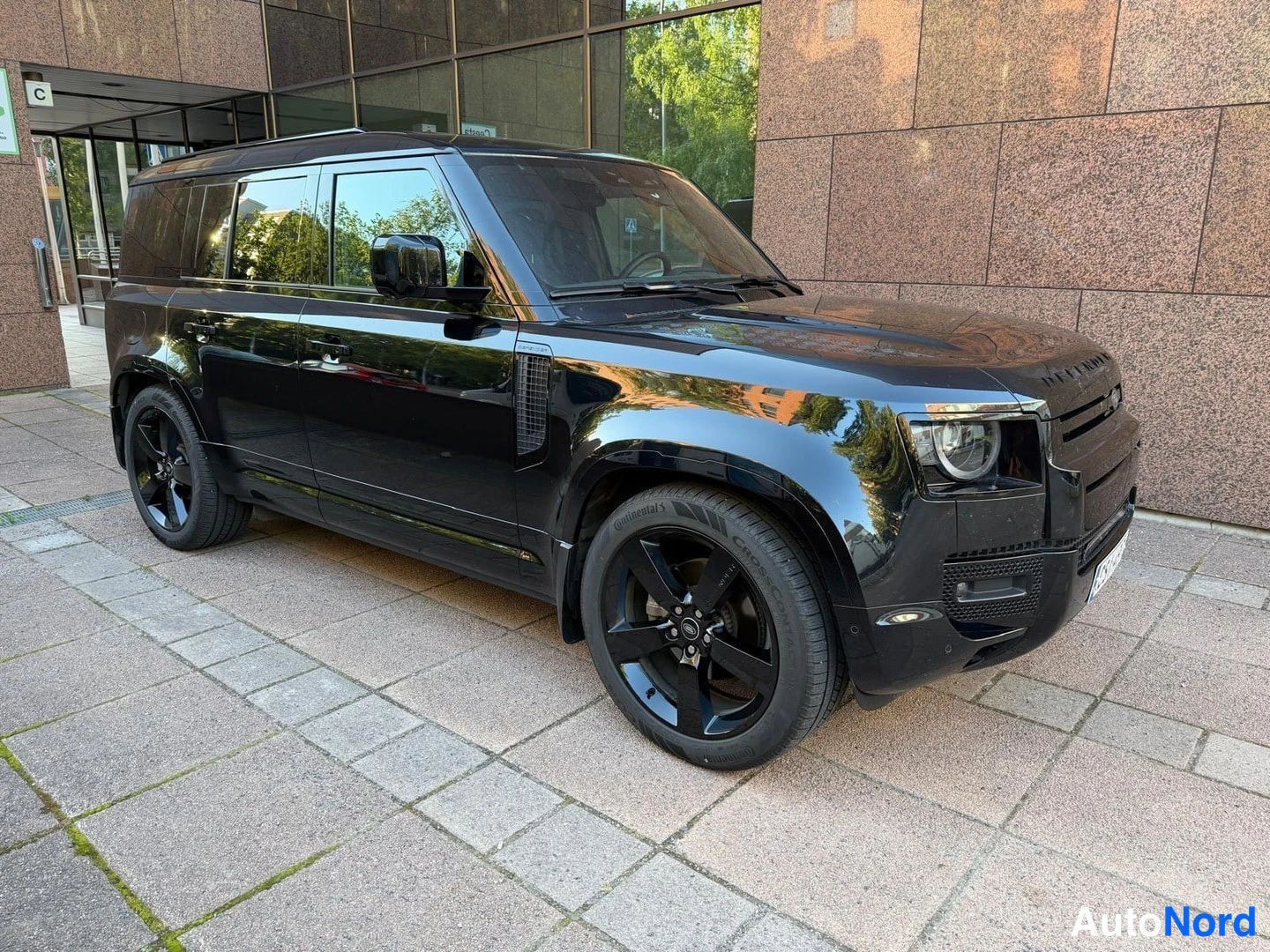 Etuviisto, nokka oikealle: Musta Land Rover Defender, kolmelisänkulmaisessa kuvassa auton keula ja vasen kylki; mustat kiintolevyt, mustat pultit ja katukuvaa kivijulkisivun edessä