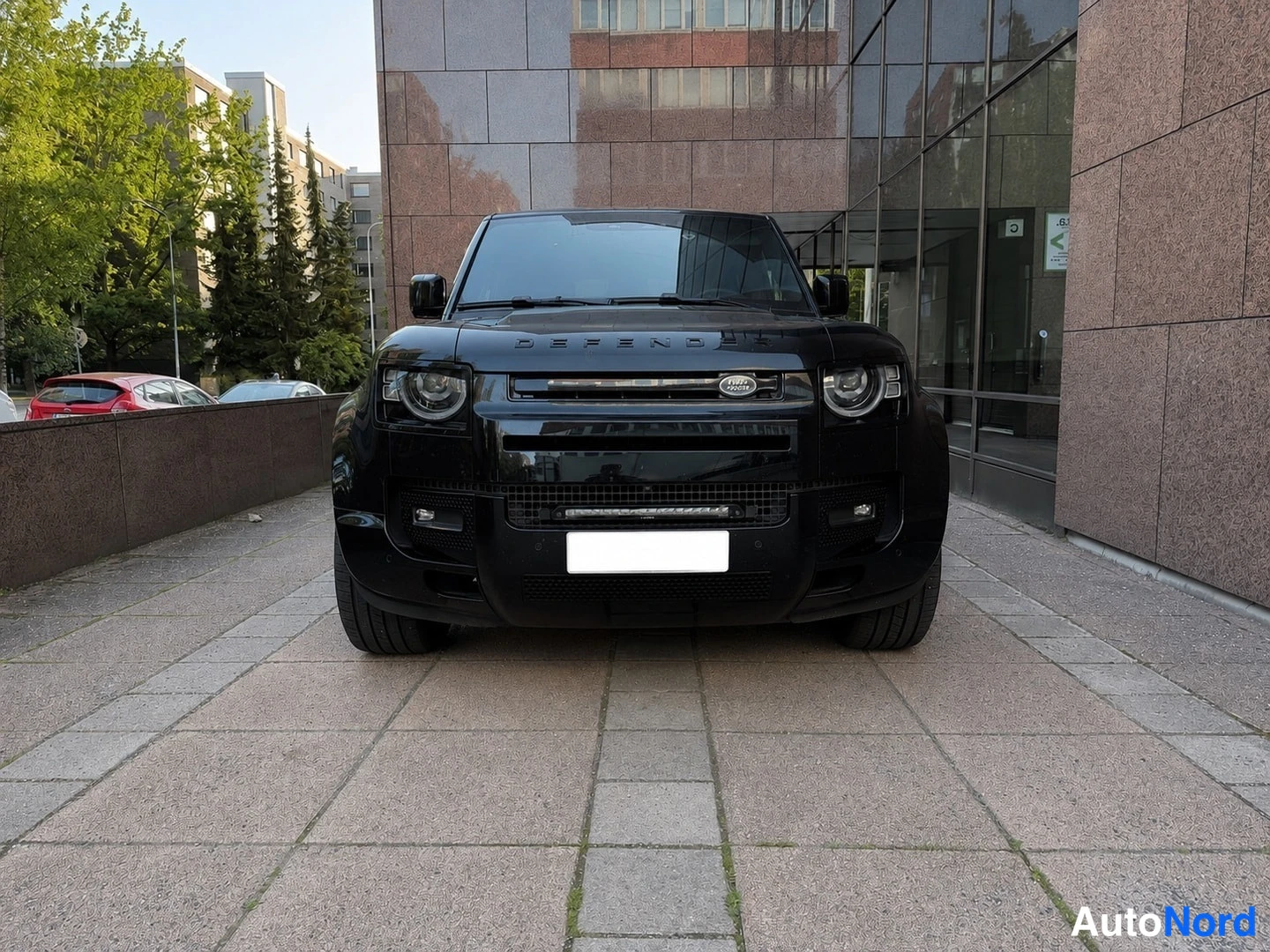 Edestä: Musta Land Rover Defender, keulakuva edestäpäin; mustat kiintolevyt, mustat pultit ja katukuvaa kivijulkisivun edessä.