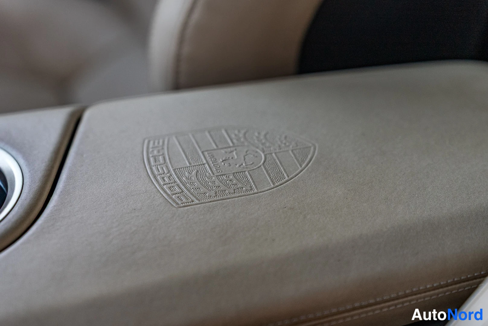 Yksityiskohta: Beige armrest with embossed Porsche crest, blurred interior in background.