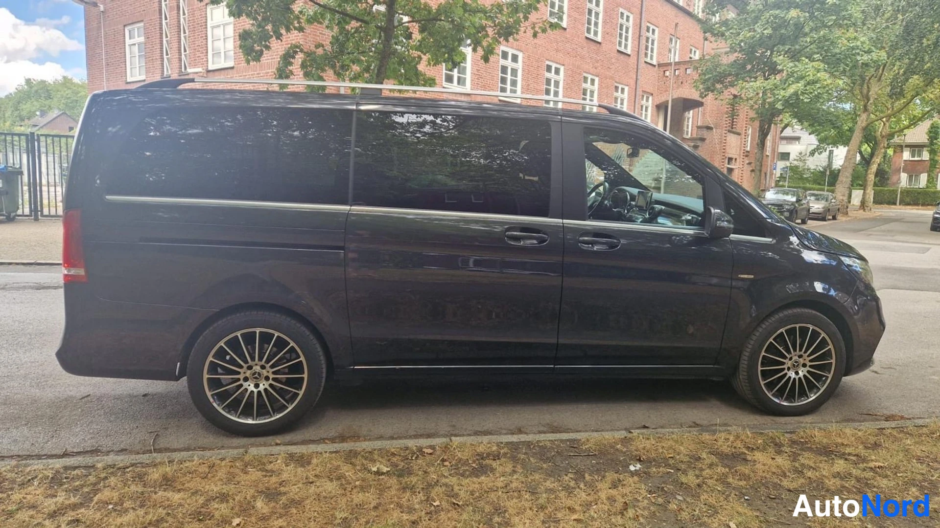 Etuviisto, nokka oikealle: Tummanharmaa pakettiauto sivulta etukulmasta oikealta, auringon heijastuksia korin sivuissa, kadun varrella puun edessä.