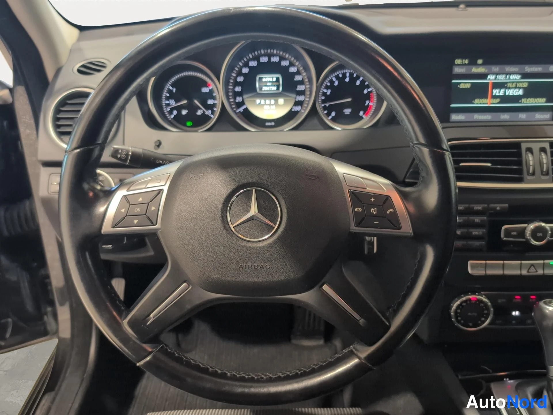 Edestä: Mercedes-Benzin ratti ja mittaristot lähikuvassa; rattitarrastissa lukee AIRBAG, taustalla infonäyttö ja radio.