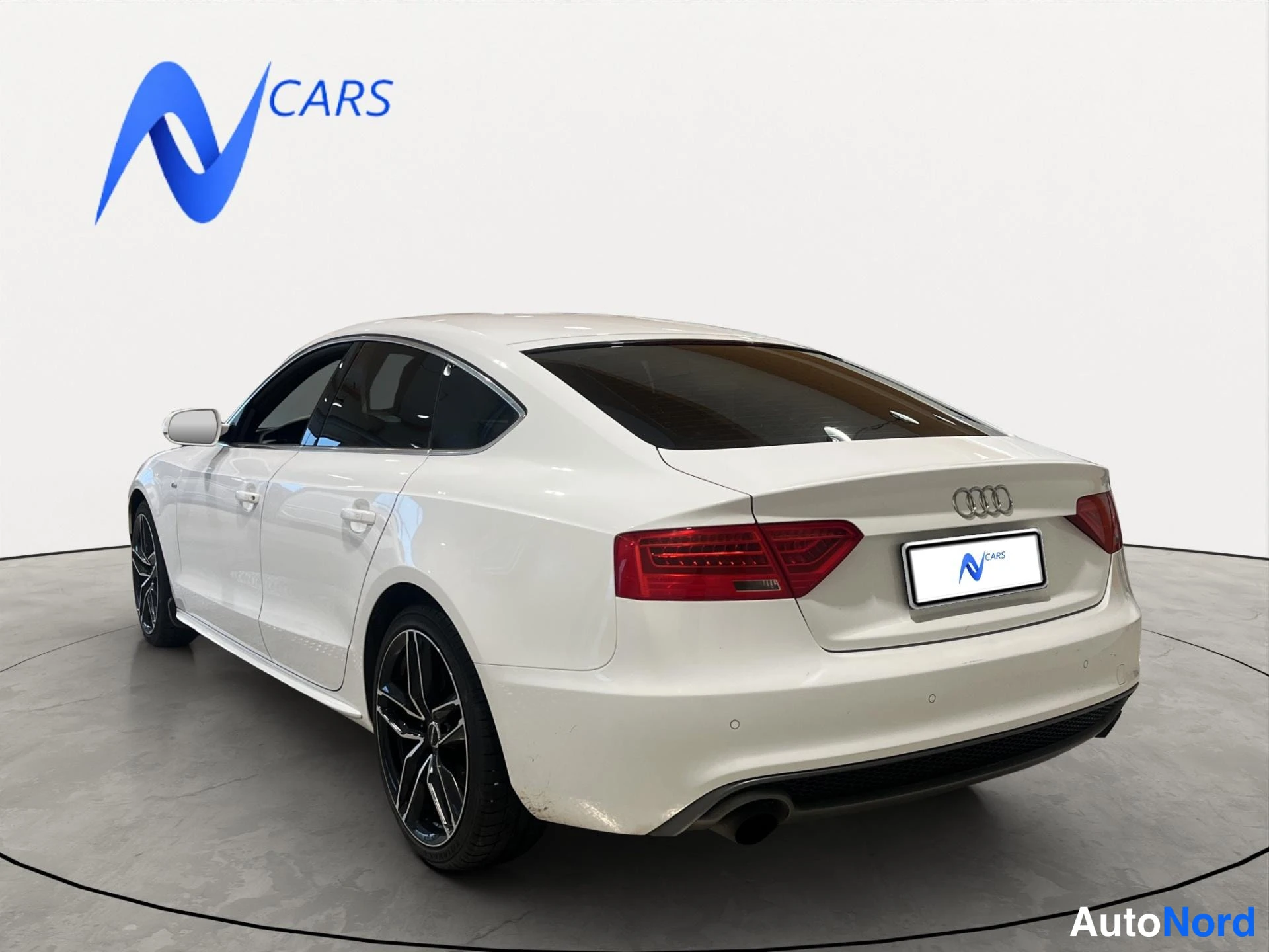Takaviisto: Valkoinen Audi A5 Sportback takakulmasta oikealta, punaiset takavalot, mustat vanteet ja kattoluukku, N Cars -logo taustalla.