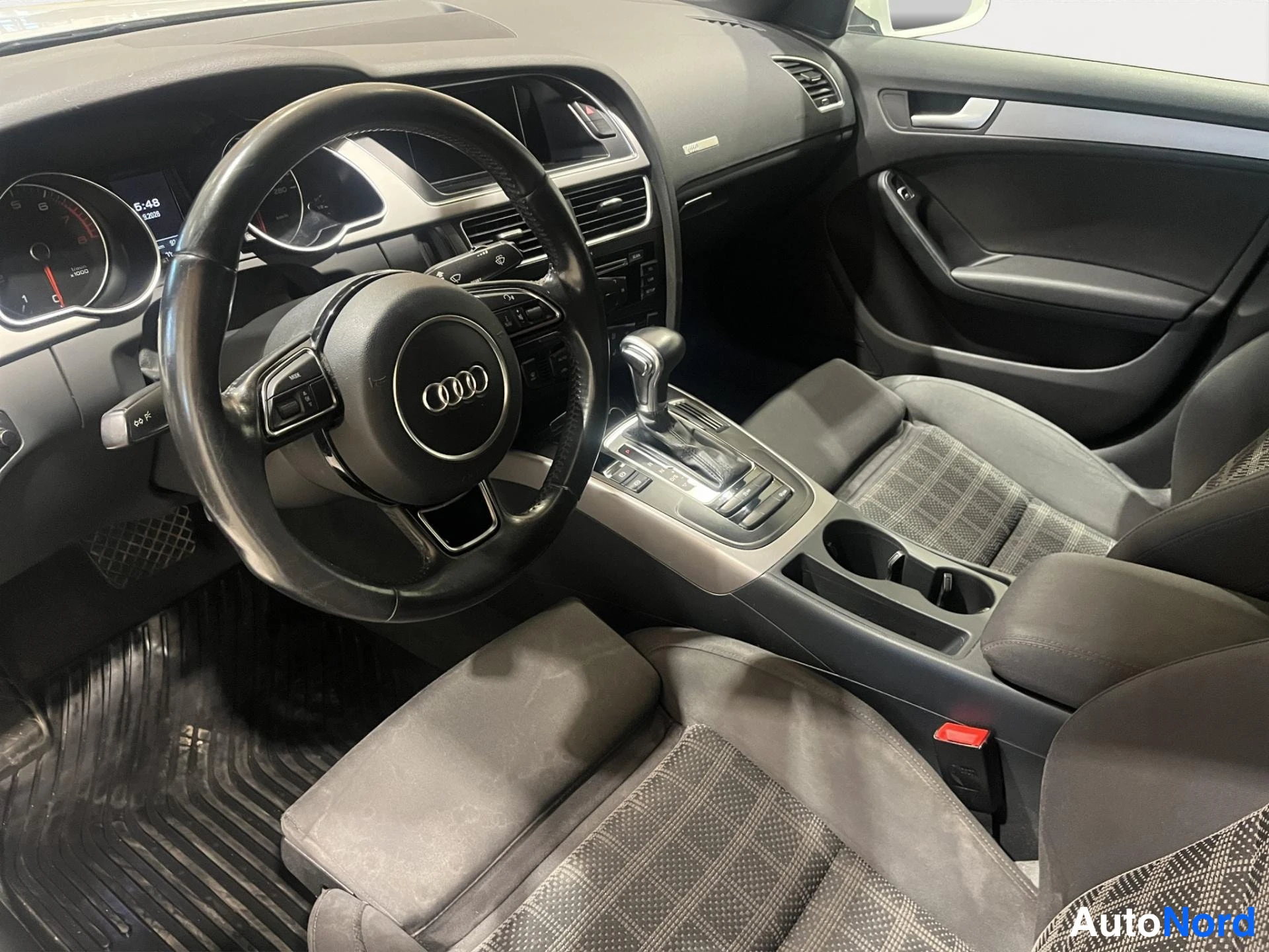 Muu: Audi-autojen sisätilat ohjaamosta nähtynä: Audi-logollinen ohjauspyörä, automaattivaihteen valitsin ja harmaat kangasistuimet.