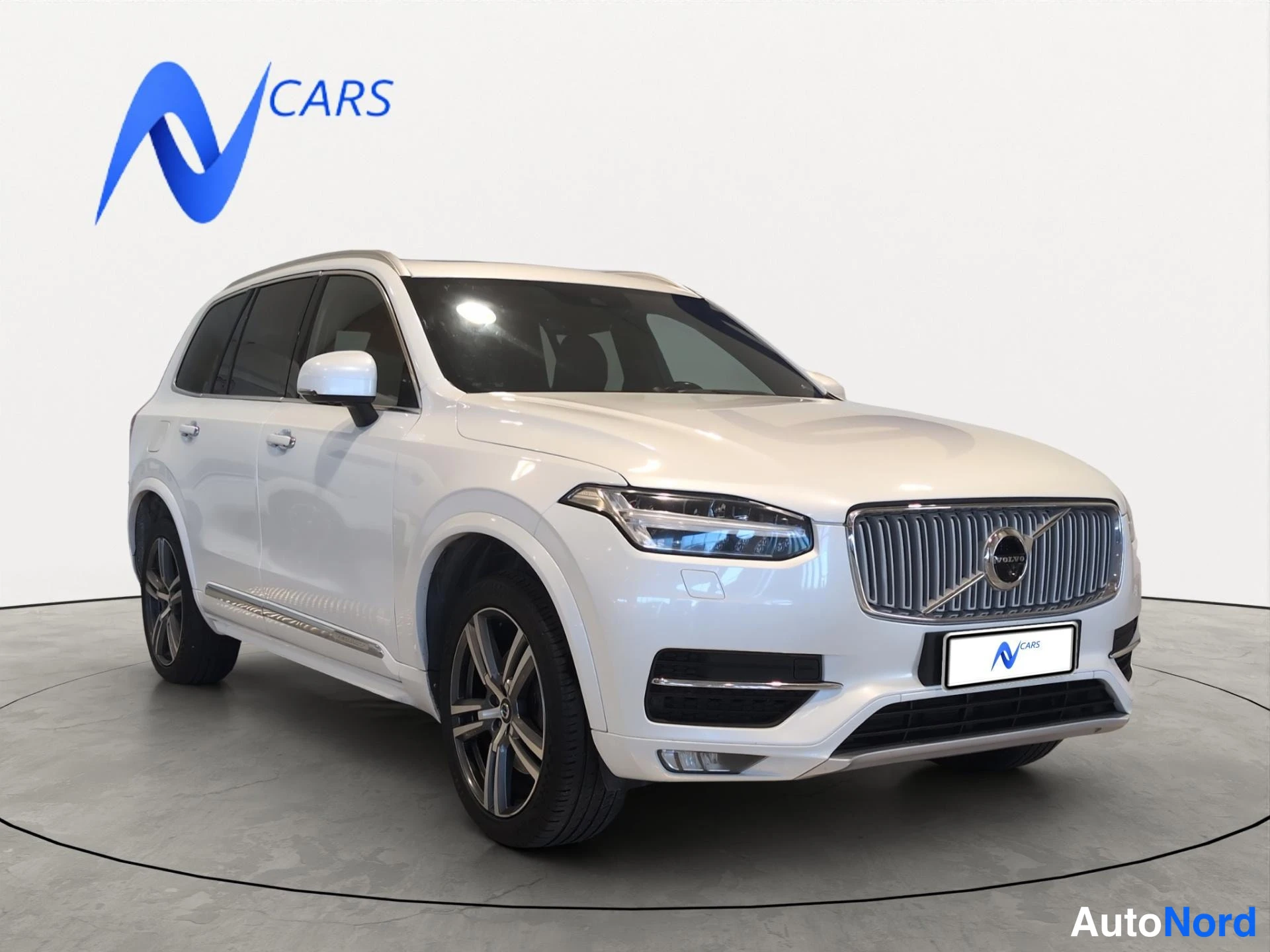 Etuviisto, nokka oikealle: Valkoinen Volvo XC90 -maasturista etuviisto oikealta; etupaneeli ja vasen kylki näkyvät studiomaisella parkettilattialla.
