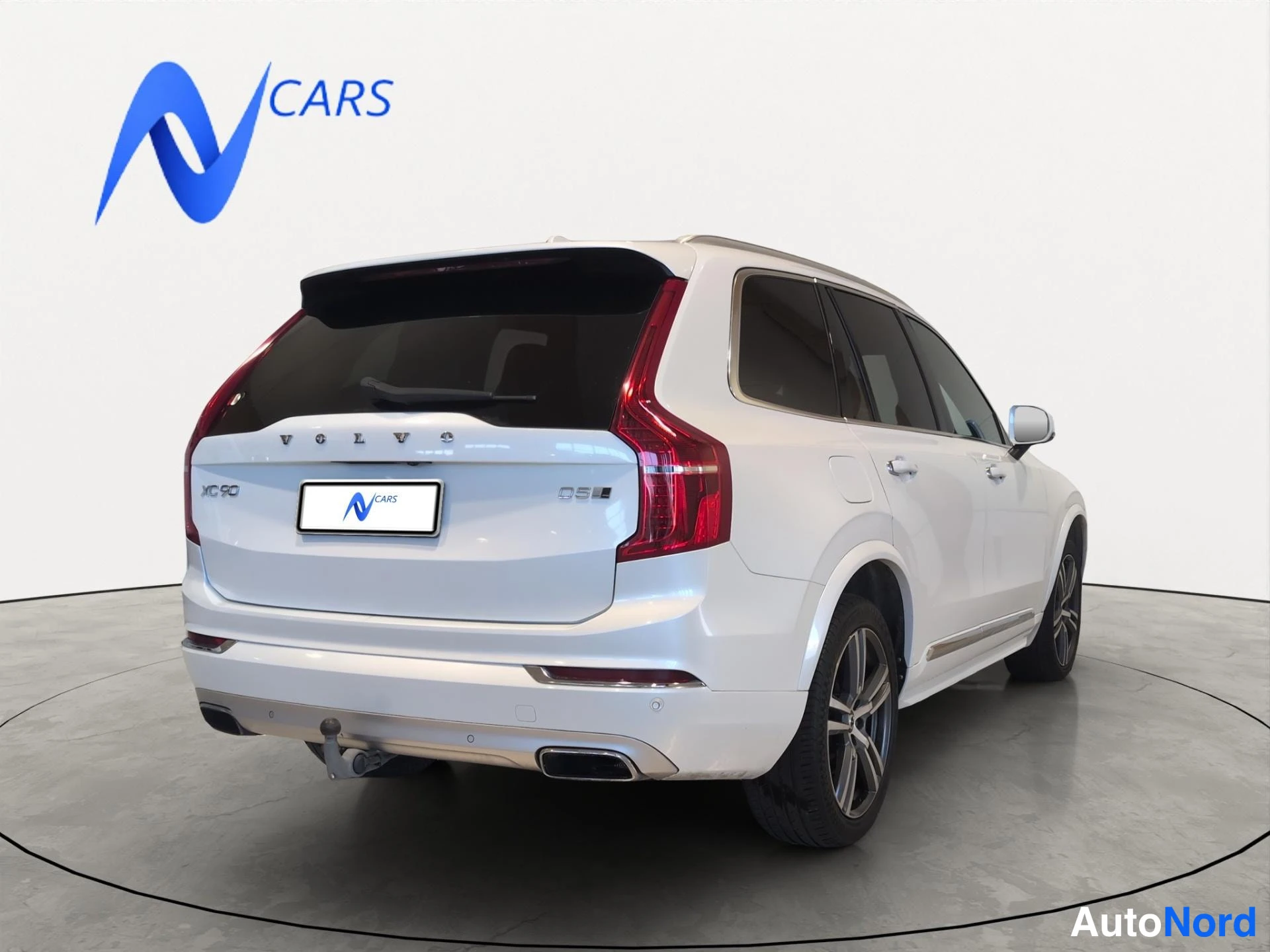 Takaviisto: Valkoisen Volvo XC90 -maasturin takaviisto vasemmalta; takavalot, XC 90- ja D5-merkinnät sekä pyykinostokoukku näkyvät.