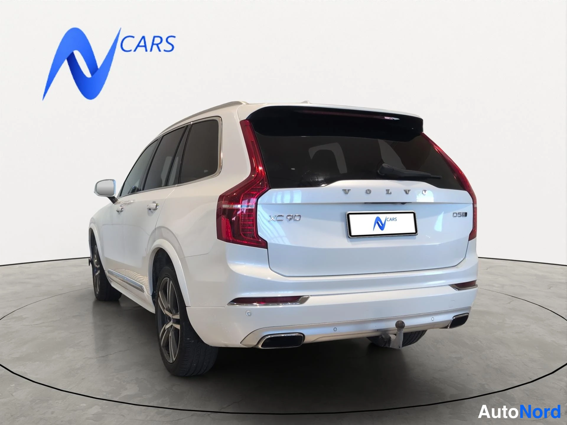 Takaviisto: Valkoisen Volvo XC90:n takaviisto oikealta; VOLVO-, XC 90- ja D5-tunukset takaovessa ja takaluukussa, aihiokanta kiinnitetty.
