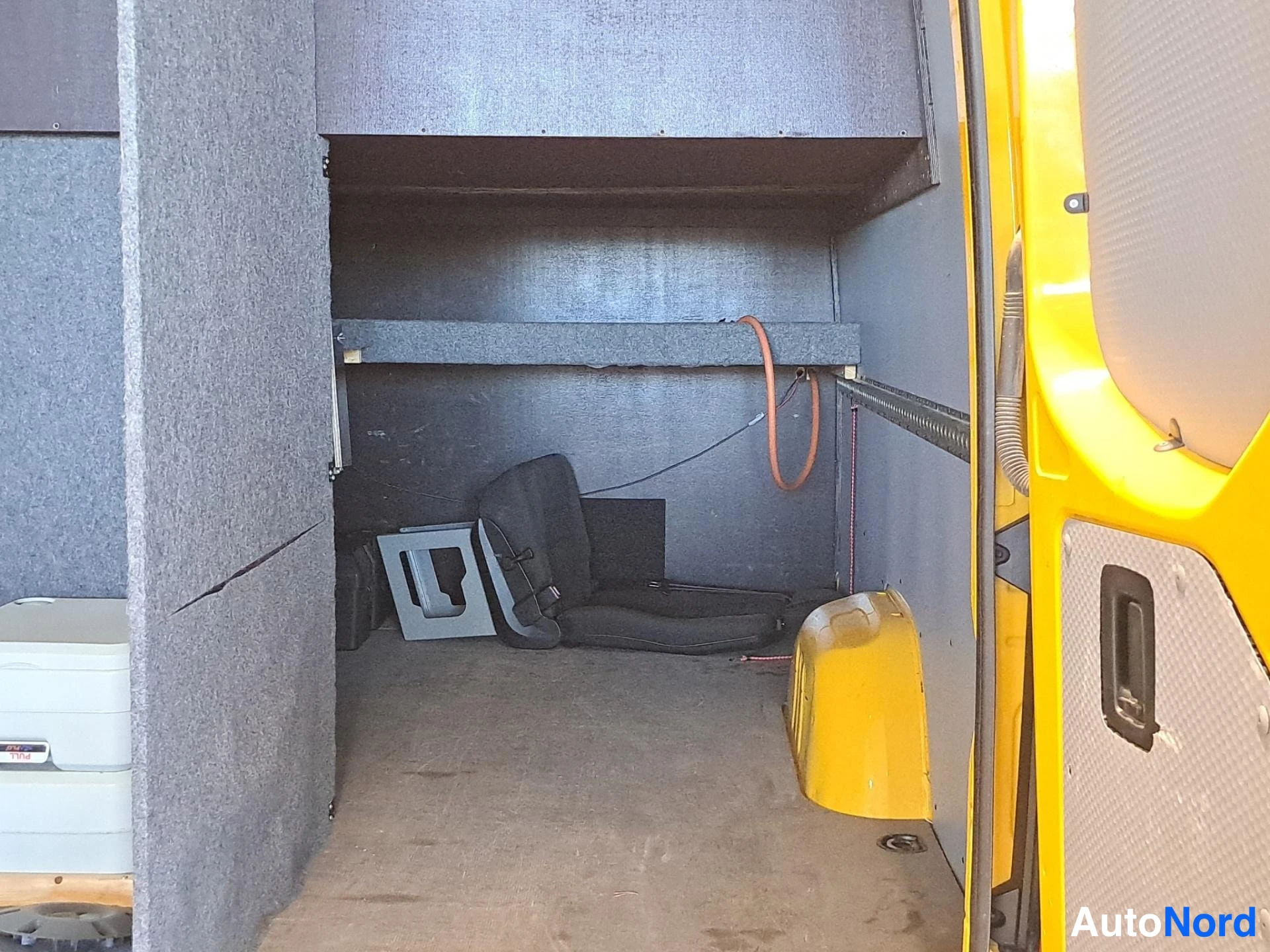 Muu: Pakettiauton tavaratila: seinät verhoiltu harmaalla huovalla, hyllytaso ja musta irtonainen istuin lattialla, keltainen siipi ja pyöräaukon suoja näkyvät oikeal