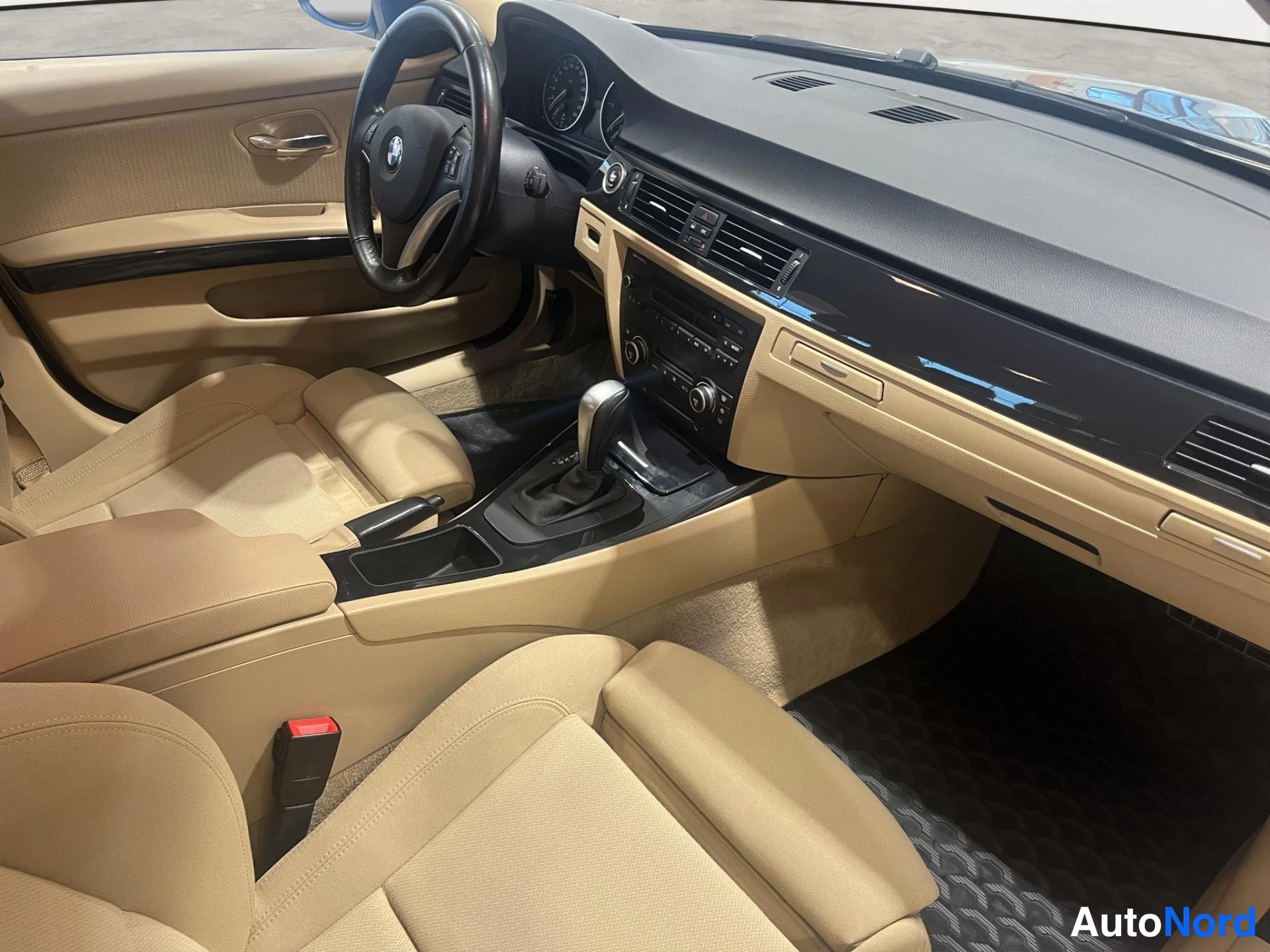 Muu: Auton sisätilat: beige verhoilu ja kojelauta, BMW-ohjauspyörä, automaattivaihteen vaihdekeila ja keskikonsolin radio näkyvät.
