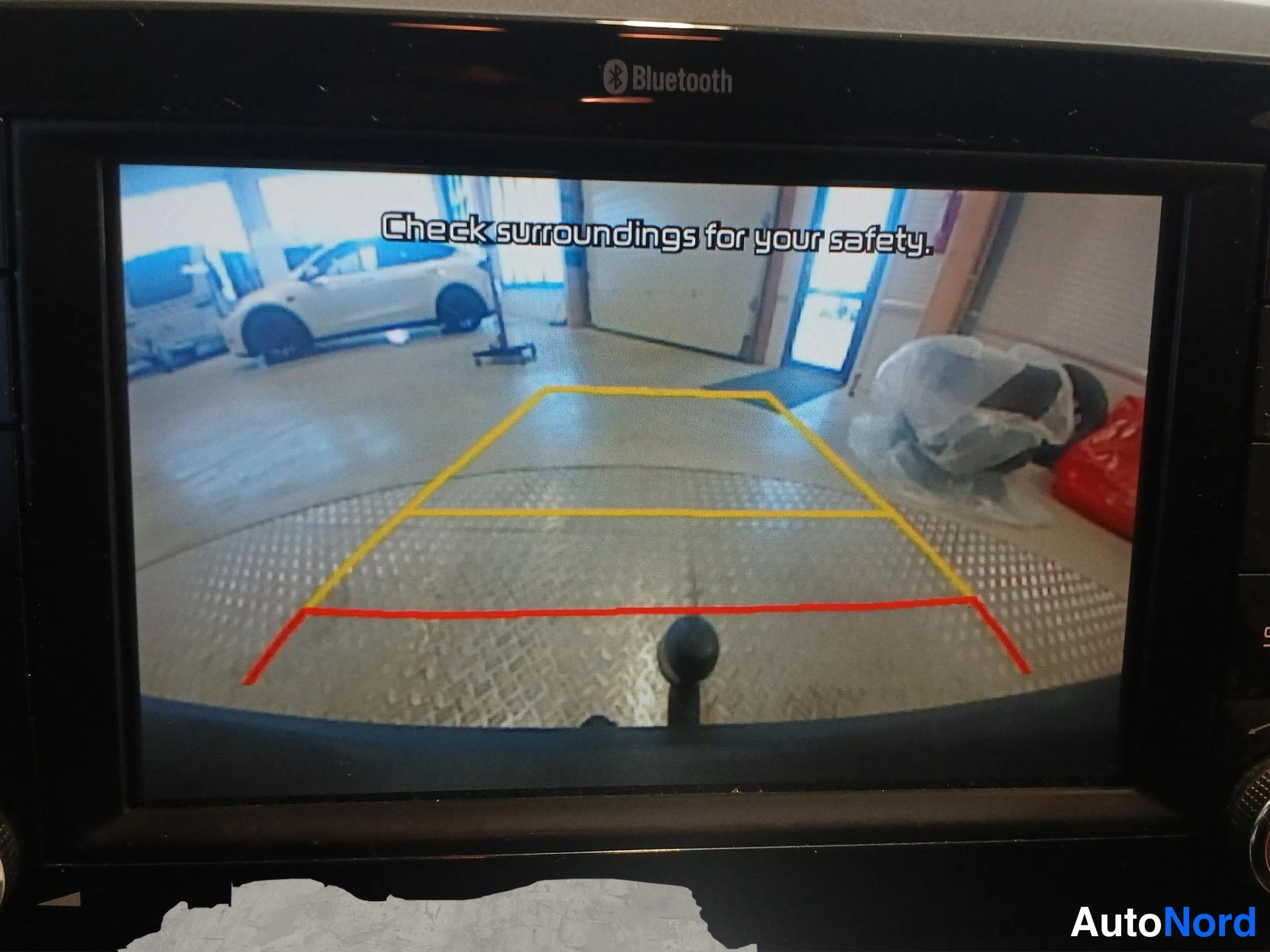 Yksityiskohta: Infotainment-näytöllä näkyy takakamera-kuva varoitusviestillä 'Check surroundings for your safety.' ja keltais-punaiset ohjaviivat.