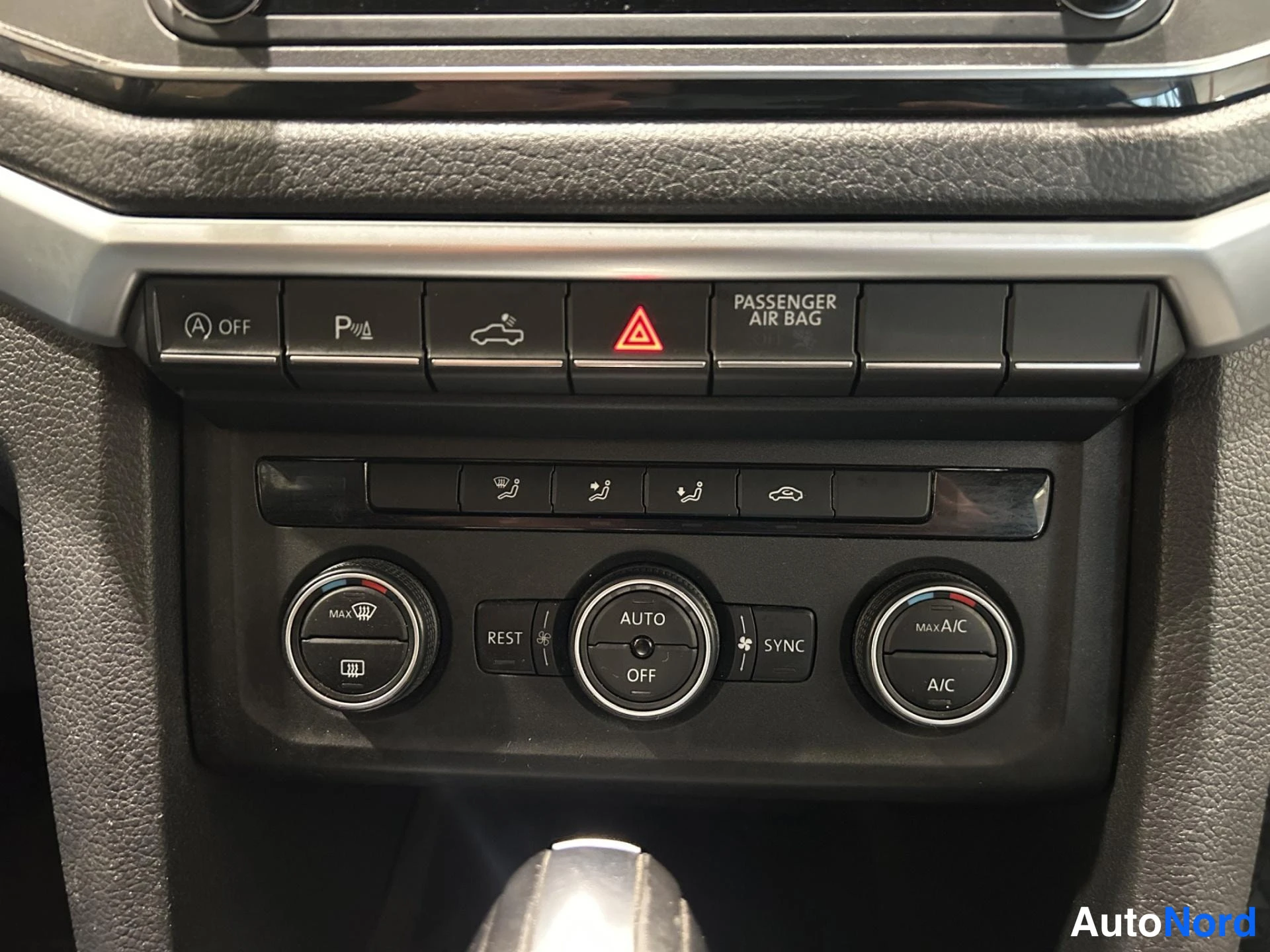Muu: Kojelaudan ilmastointipaneelin lähikuva: nappi AUTO, OFF, SYNC, A/C ja MAX A/C, sekä ylimmällä rivilla parkkitutkan P-nappi, varoitusvalo ja PASSANGER AIR BAG -