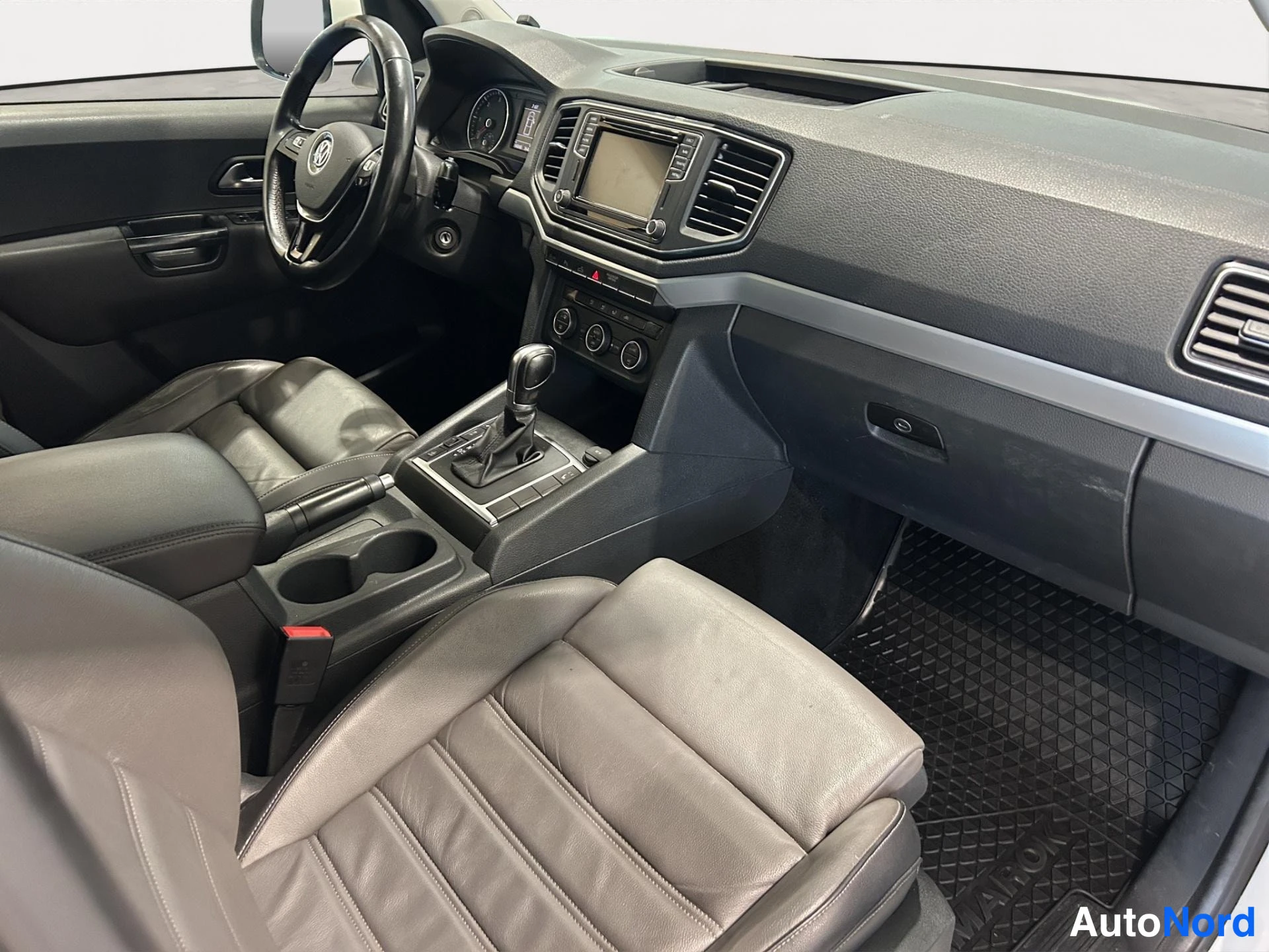 Muu: Amarok-mallin ohjaamo etuistuimilta kuvattuna: Volkswagen-ohjauspyörä, automaattivaihteen valitsin, radio ja tummat nahkaiset etuistuimet.