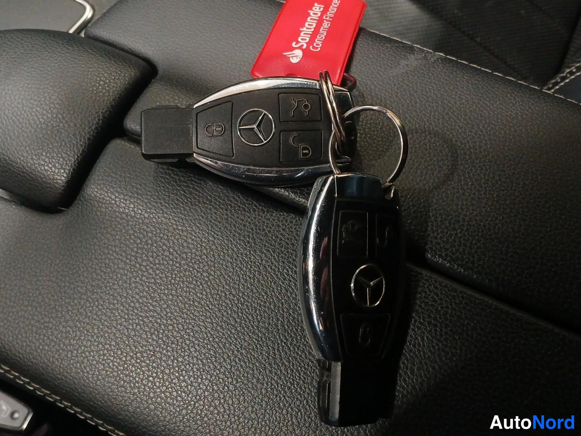 Muu: Kaksi Mercedes-Benzin avainetäisyyssäädintä mustassa nahkapinnassa, mukana punainen Santander Consumer Finance -avainkilpi.