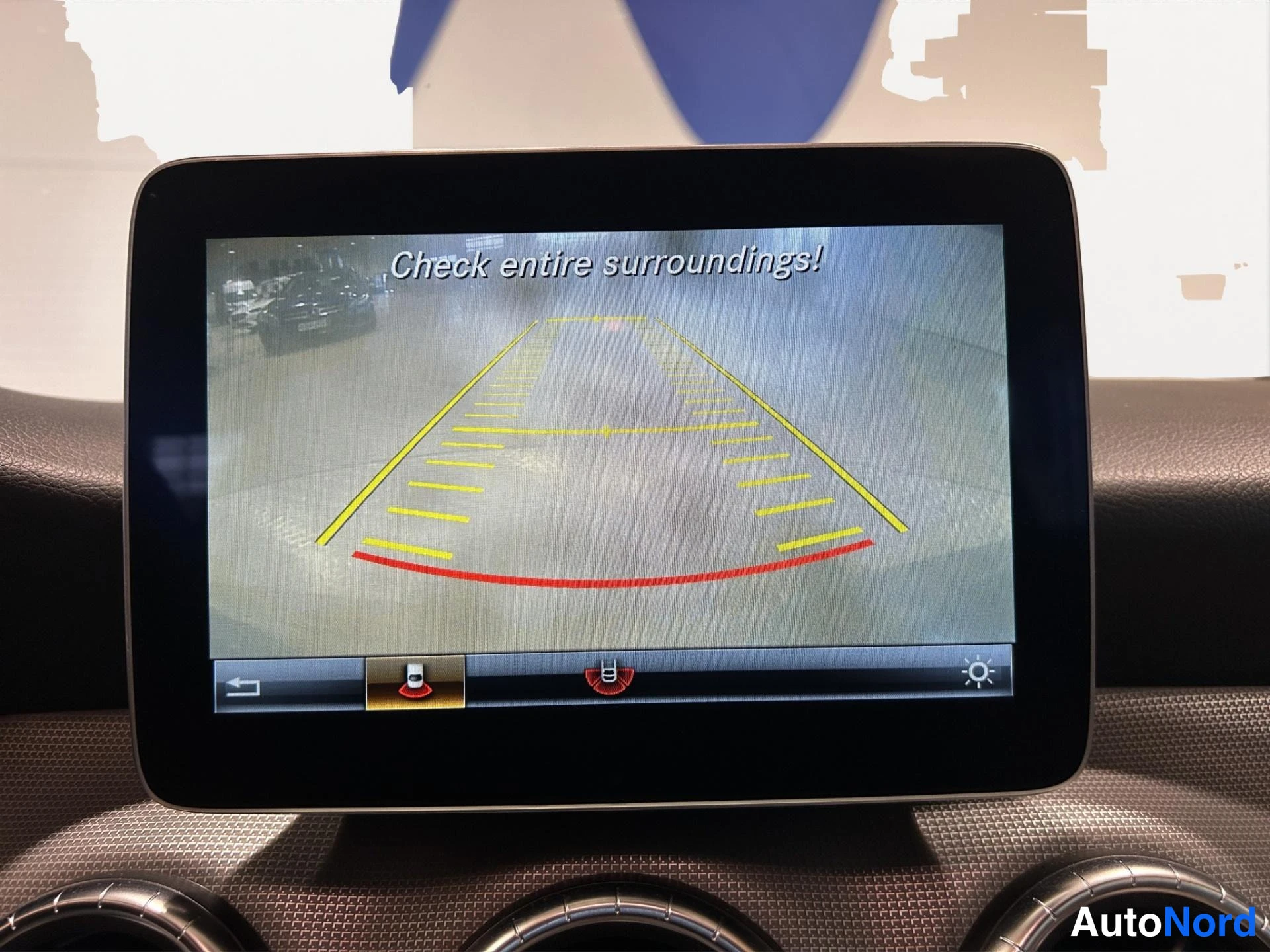 Yksityiskohta: Mercedes-Benzin näyttö, jossa takana ajokamera: teksti 'Check entire surroundings!', keltainen ja punainen kaistaneuvonta ruudussa, kuvakepalkki alareunassa.