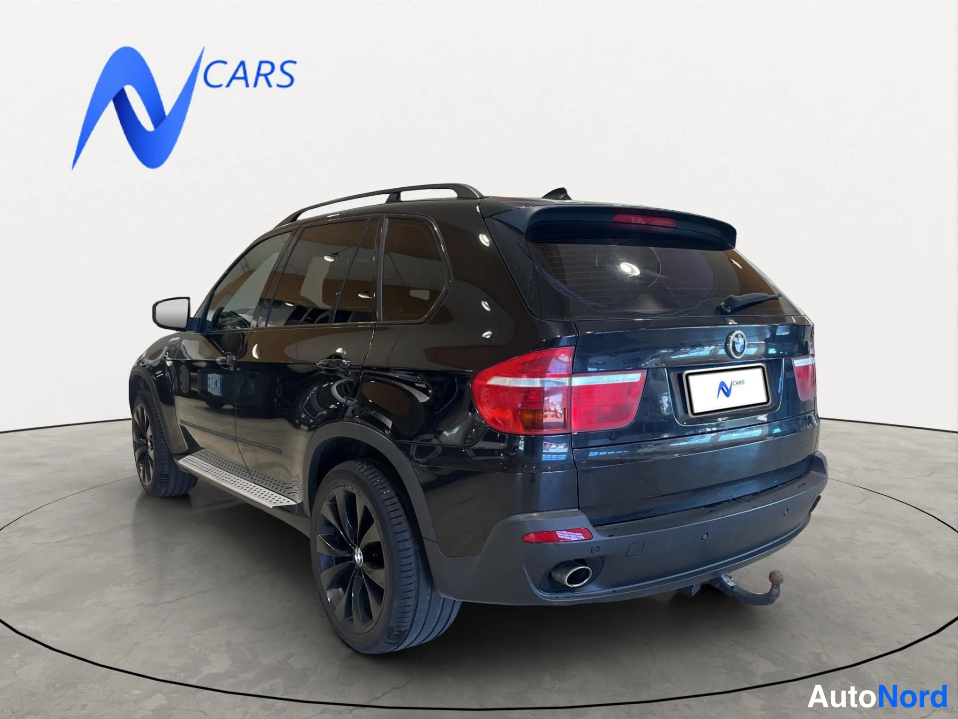 Takaviisto: Musta BMW X5 takakulmasta vasemmalta, kattotelineet, vetokoukku ja mustat vanteet näkyvät.