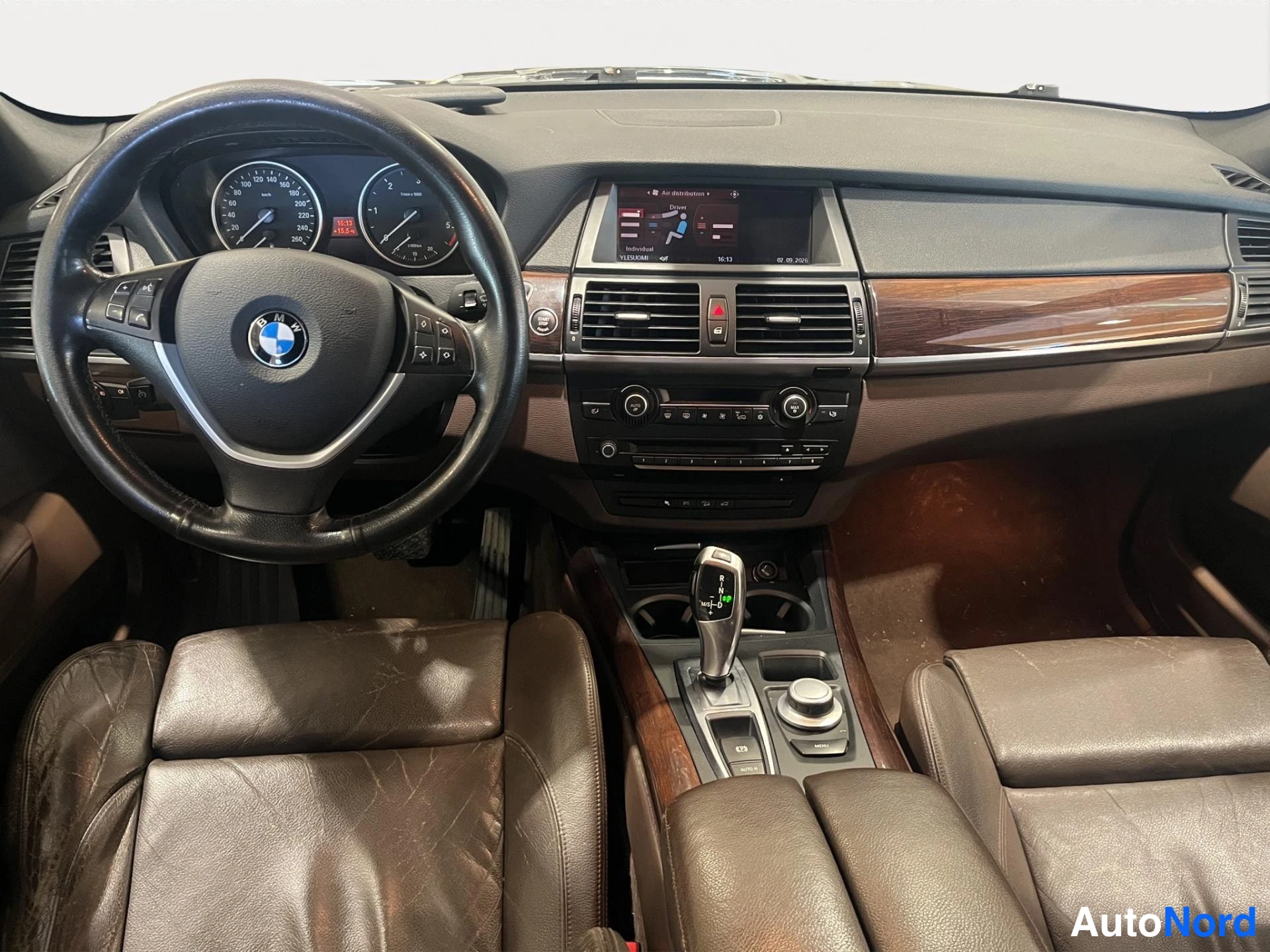 Muu: Ohjaamo suoraan edestä: BMW-ohjauspyörä, mittarit, keskinäytön infotainment-järjestelmä päällä, ruskeat nahkaistuimet ja automaattivaihteen valitsin.