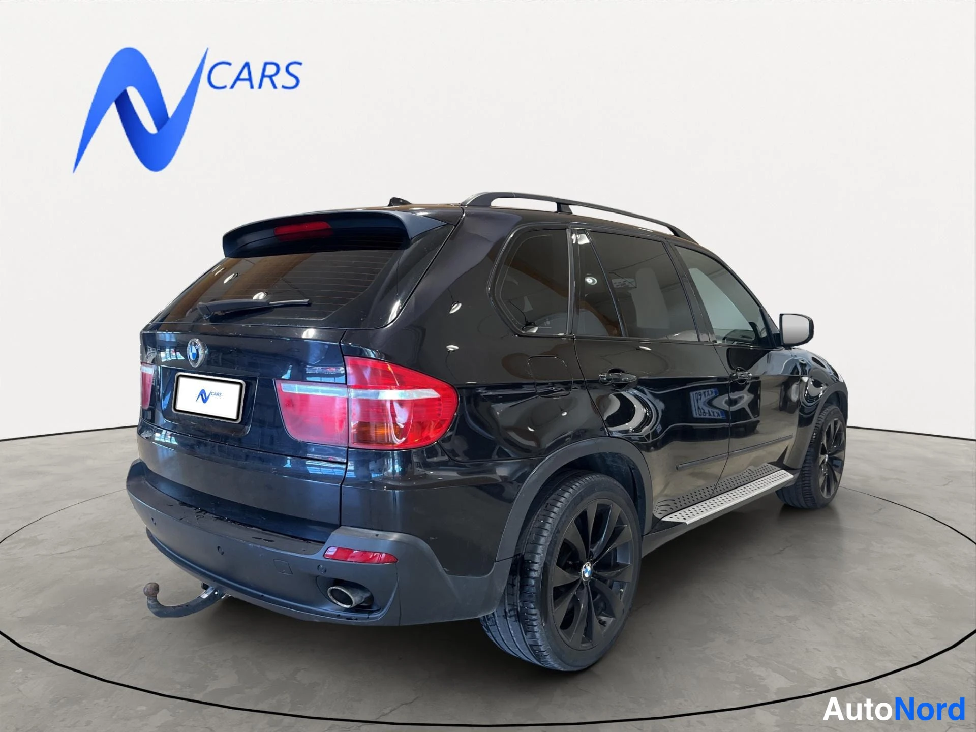 Takaviisto: Musta BMW X5 takakulmasta oikealta, kattotelineet, kylkiaskelmat ja vetokoukku näkyvät studiotaustaa vasten.