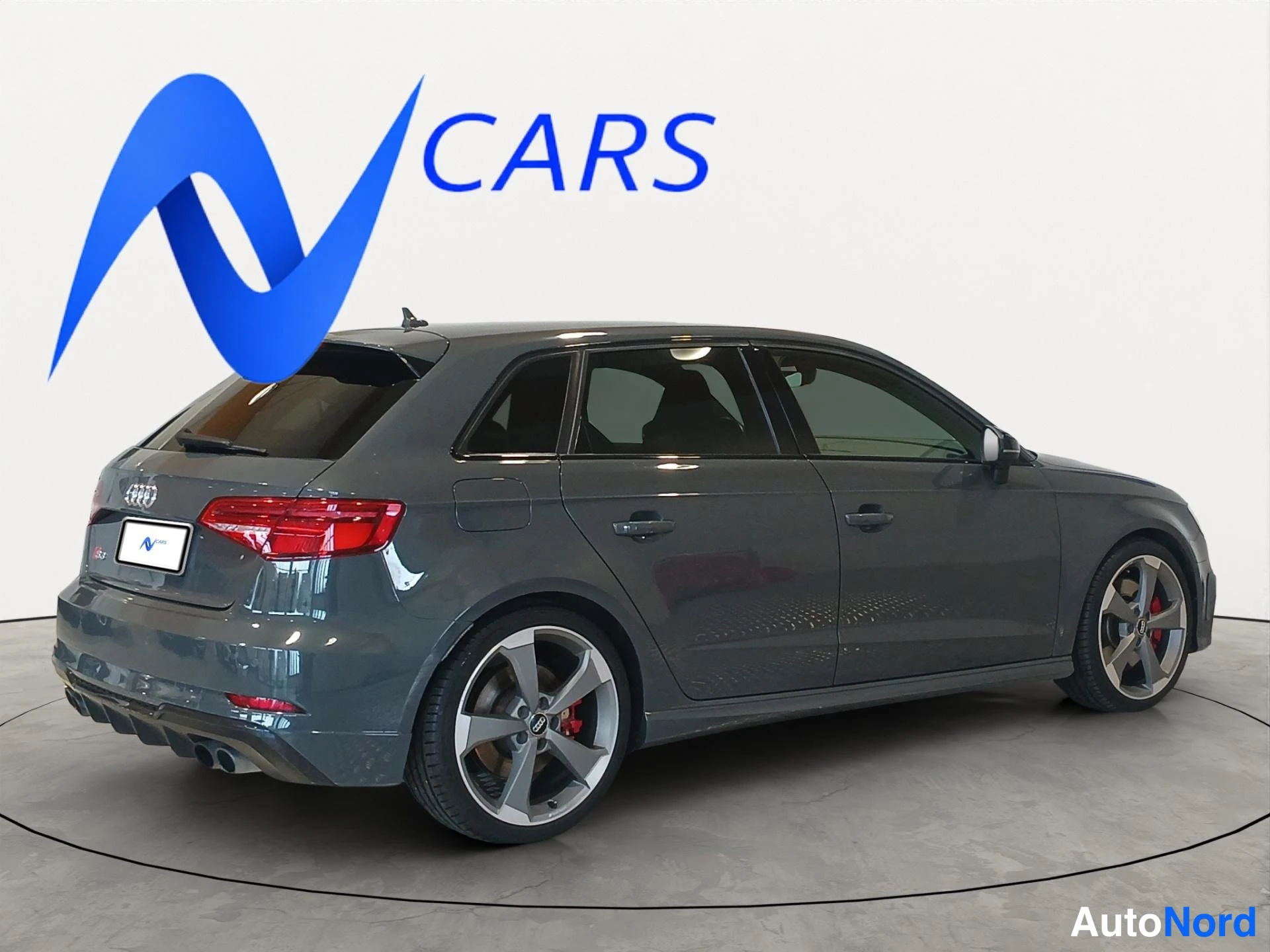 Takaviisto: Tummanharmaa Audi A3 Sportback takavasemmasta kulmasta kuvattuna; näkyvissä takavalot, takaluukki, Audi-merkki, Audi-laatikkomerkki vanteissa ja punainen jarrus