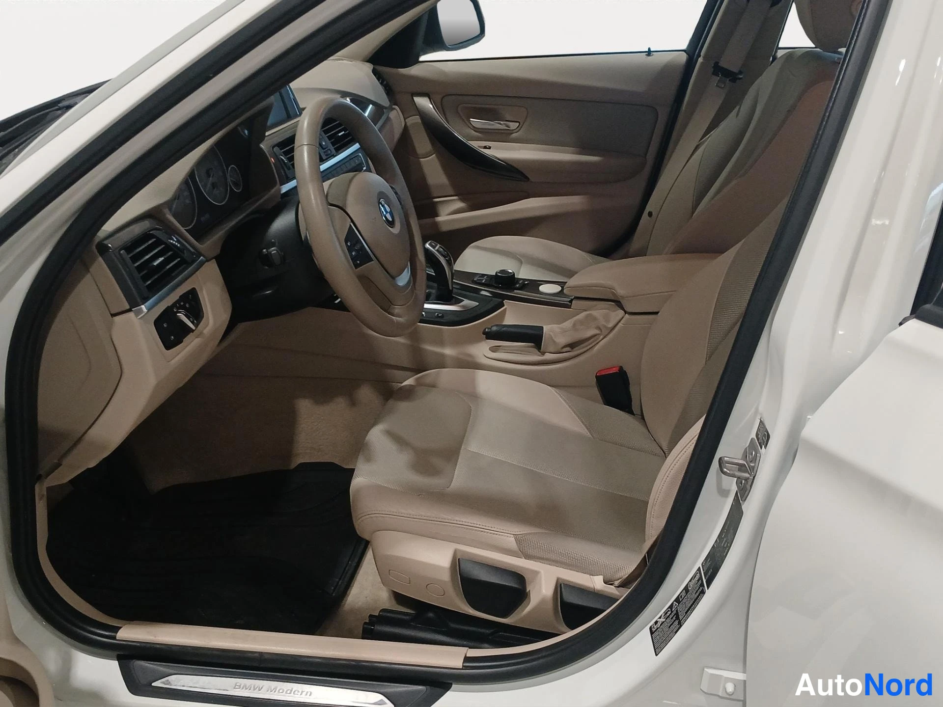 Muu: BMW:n etuistuimin sisäkuva kuljettajan puolelta: ohjauspyörä, kojelauta ja beige kankaanverhoitus, kynnyspiirtokirjoitus BMW Modern.