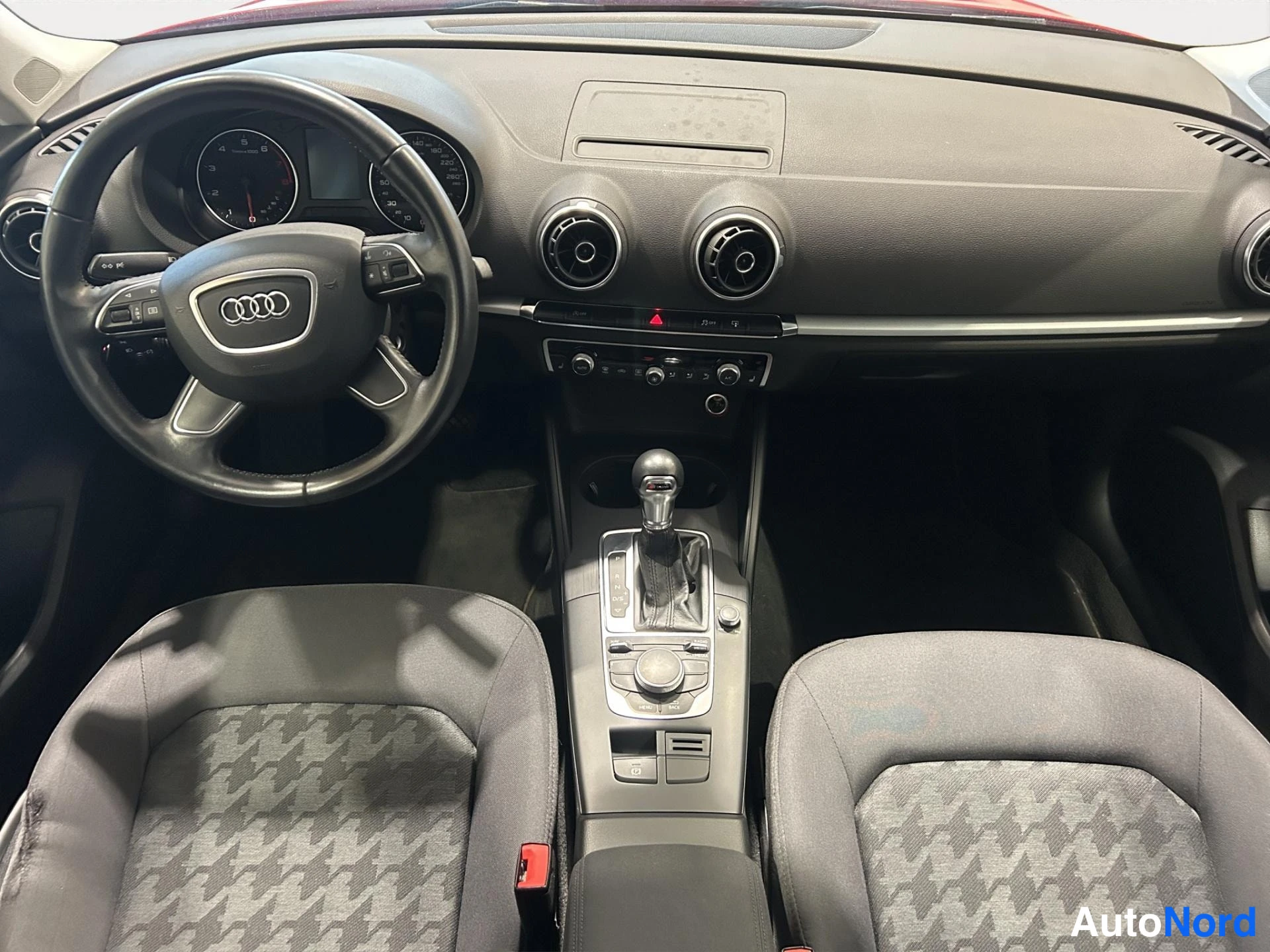 Muu: Auton sisätila: ohjauspyörä Audi-merkillä, kojetaulu ja manuaali-automaattivaihteen keila, kaksi kangasistuinta etualalla.