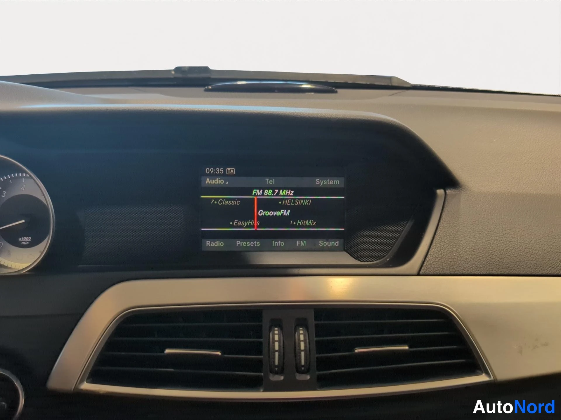 Muu: Mercedes-auton infotainment-näyttö, jossa näkyy FM 88.7 MHz radio ('GrooveFM') sekä vasemmalla osa nopeusmittarista; alaosassa ilmastointisuulake.