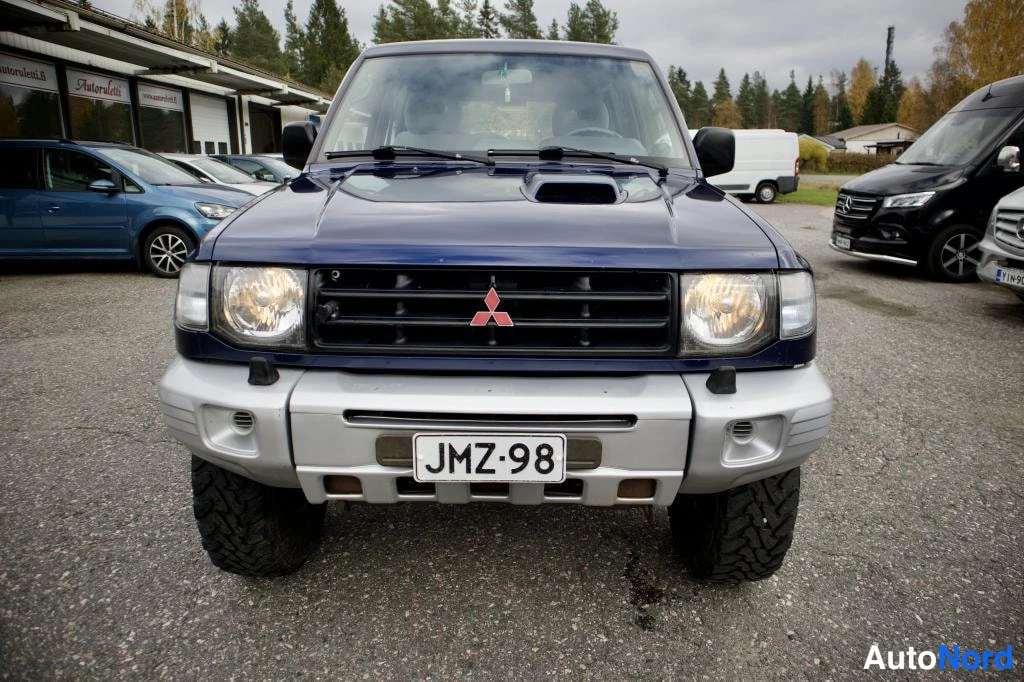 Edestä: Sinisen Mitsubishi Pajeron etunäkymä, Mitsubishin merkki säleikössä ja rekisterikilpi etupuskurissa.