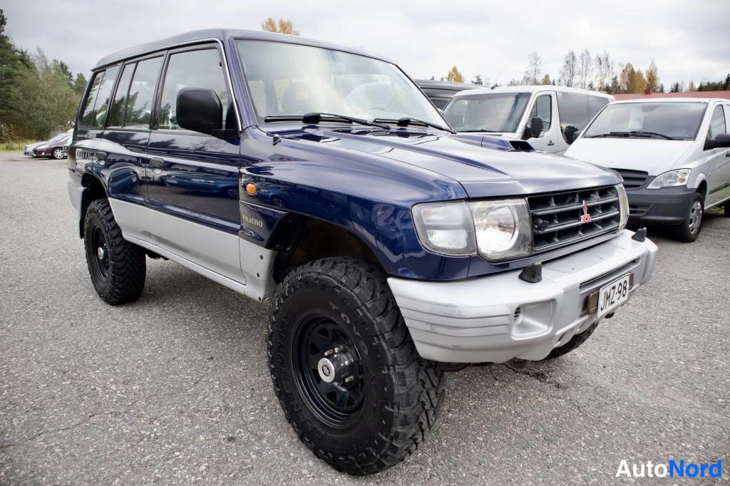 Etuviisto, nokka oikealle: Tummansininen Mitsubishi Pajero kolmeneljäsosan kulmakuvassa edessä oikealle, mustat maastorenkaat ja hopeiset alakanneluet.
