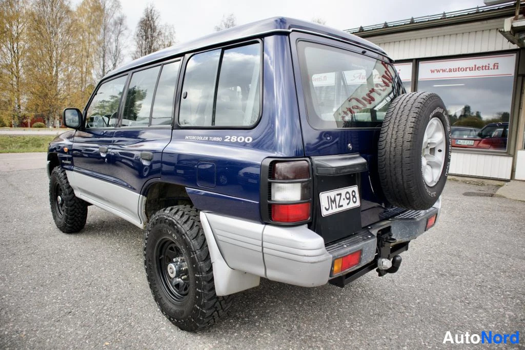 Takaviisto: Sininen Mitsubishi Pajero takaviistosta vasemmalle, varapyörä rekisterikilpi.