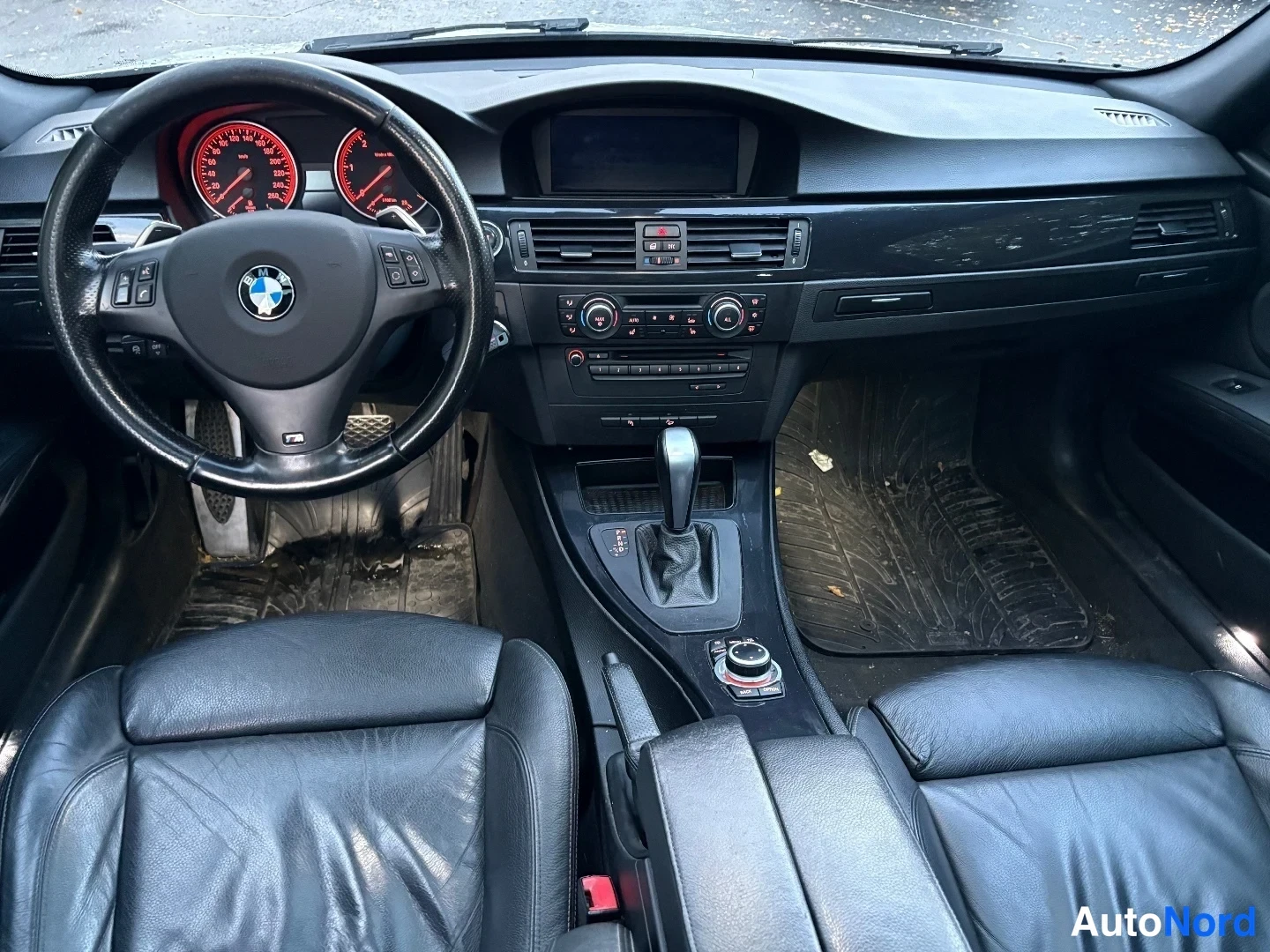 Yksityiskohta: Ohjaamo suoraan edestä kuvattuna: BMW-ohjauspyörä, punaisin valaistut mittarit, näyttö keskellä kojelautaa ja automaattivaihteen valitsin.