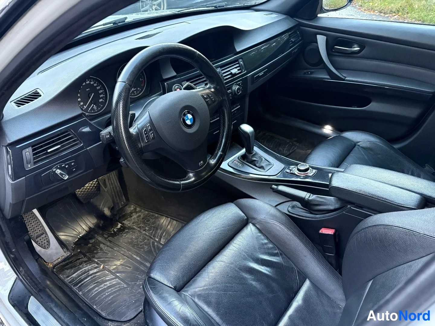 Yksityiskohta: Ohjaamo kuvattuna kuljettajan oven puolelta:BMW-ohjauspyörä, analogiset mittarit, automaattivaihteen valitsin ja mustat nahkaistuimet.
