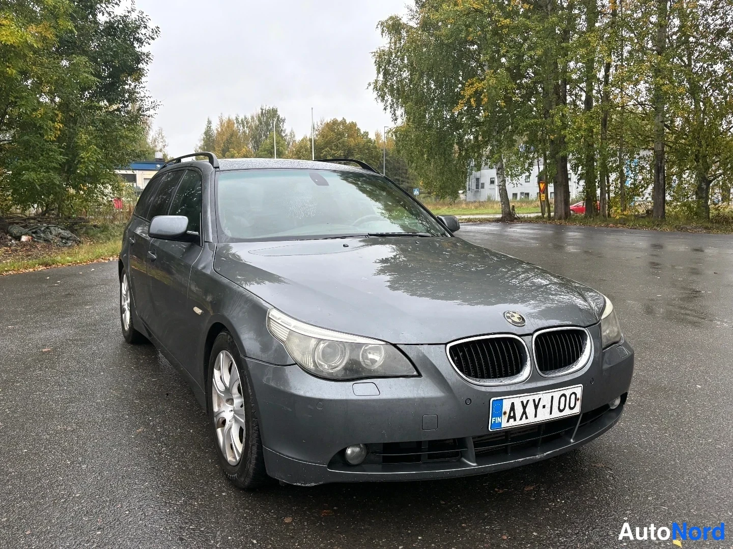 Etuviisto, nokka oikealle: Tummanharmaan BMW-farmariauton etukulmakuva oikealta; etusäleikkö, etuvalot ja kylki näkyvät sadekelin märällä parkkipaikalla. Rekisterikilpi.