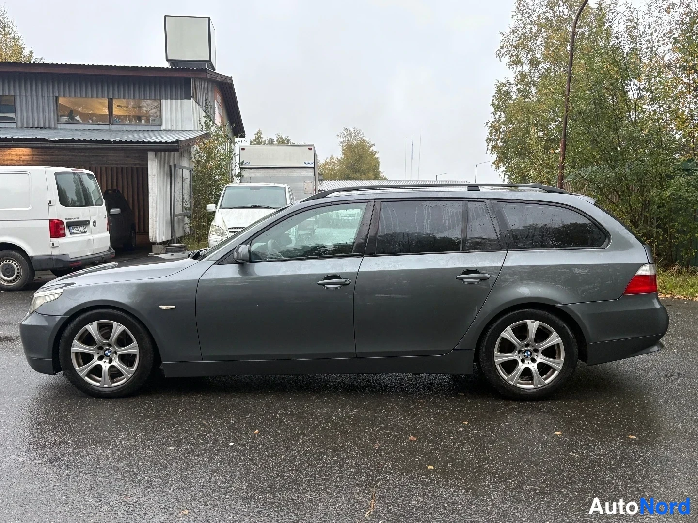 Sivulta: Tummanharmaan BMW-farmariauton sivukuva vasemmalta; ovien kahvat, kattokaiteet ja kevytmetallivanteet näkyvät asfaltoidulla pihalla.