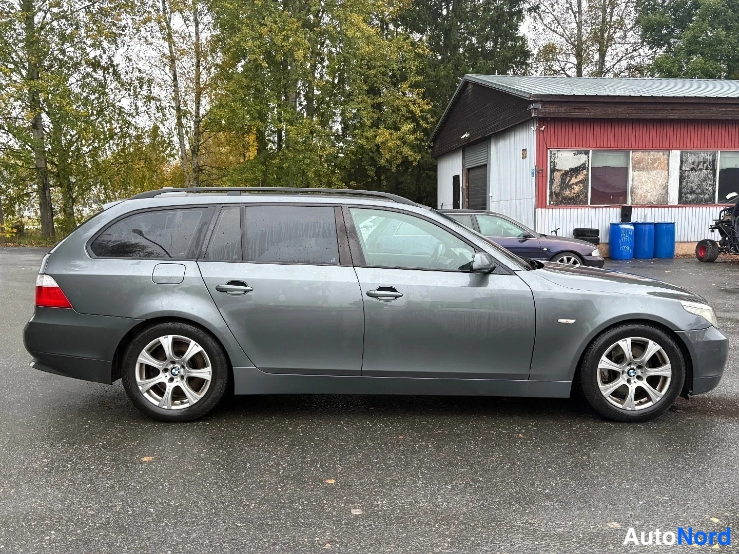 Sivulta: BMW 5-sarjan farmari sivulta kuvattuna, tummanharmaa kori, katuhälyt ja alumiinivanteet näkyvissä märällä pihalla.
