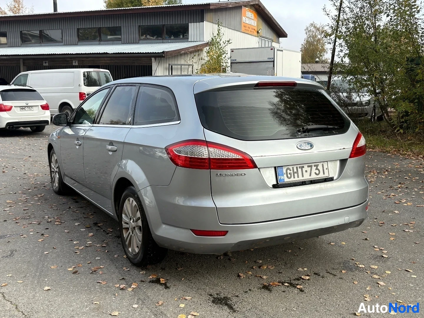 Takaviisto: Ford Mondeo Farmari takaa viistosti kuvattuna, hopeametallinen korimalli pihamaalla muiden ajoneuvojen joukossa.