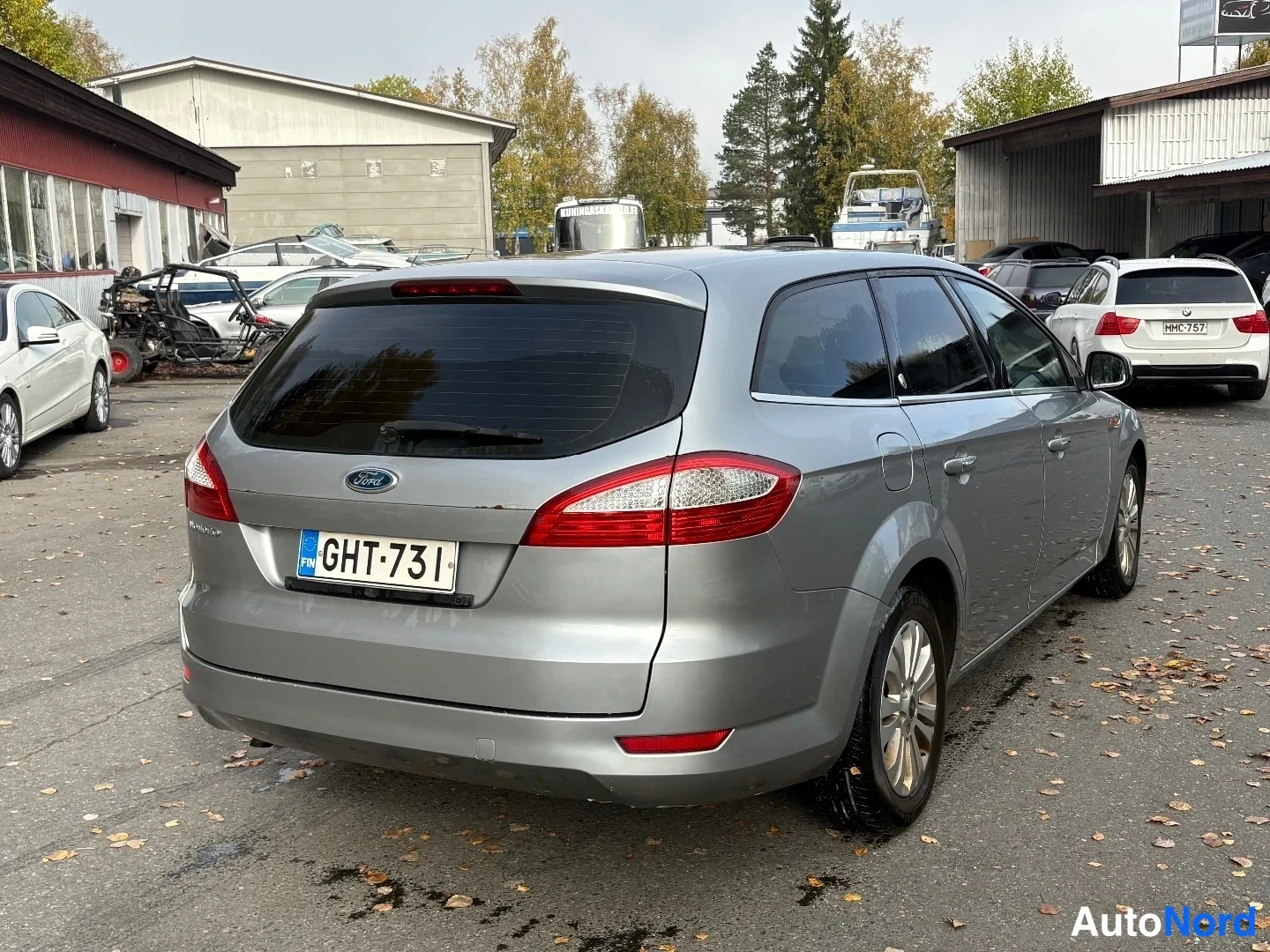 Takaviisto: Ford Mondeo Farmari takaa ja vasemmalta sivulta kuvattuna, hopeametallinen korimalli pihamaalla muiden ajoneuvojen joukossa.