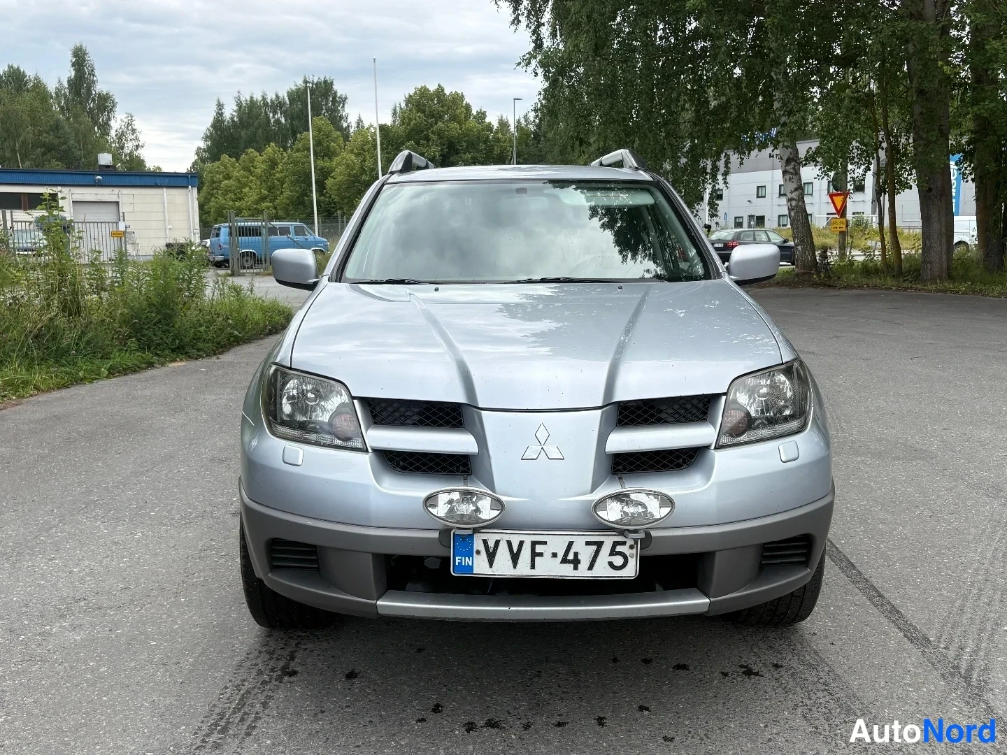 Edestä: Hopeanvärinen Mitsubishi Outlander suoraan edestäpäin, Mitsubishi-tähti ja rekisteritunnus, lisävalot puskurissa.