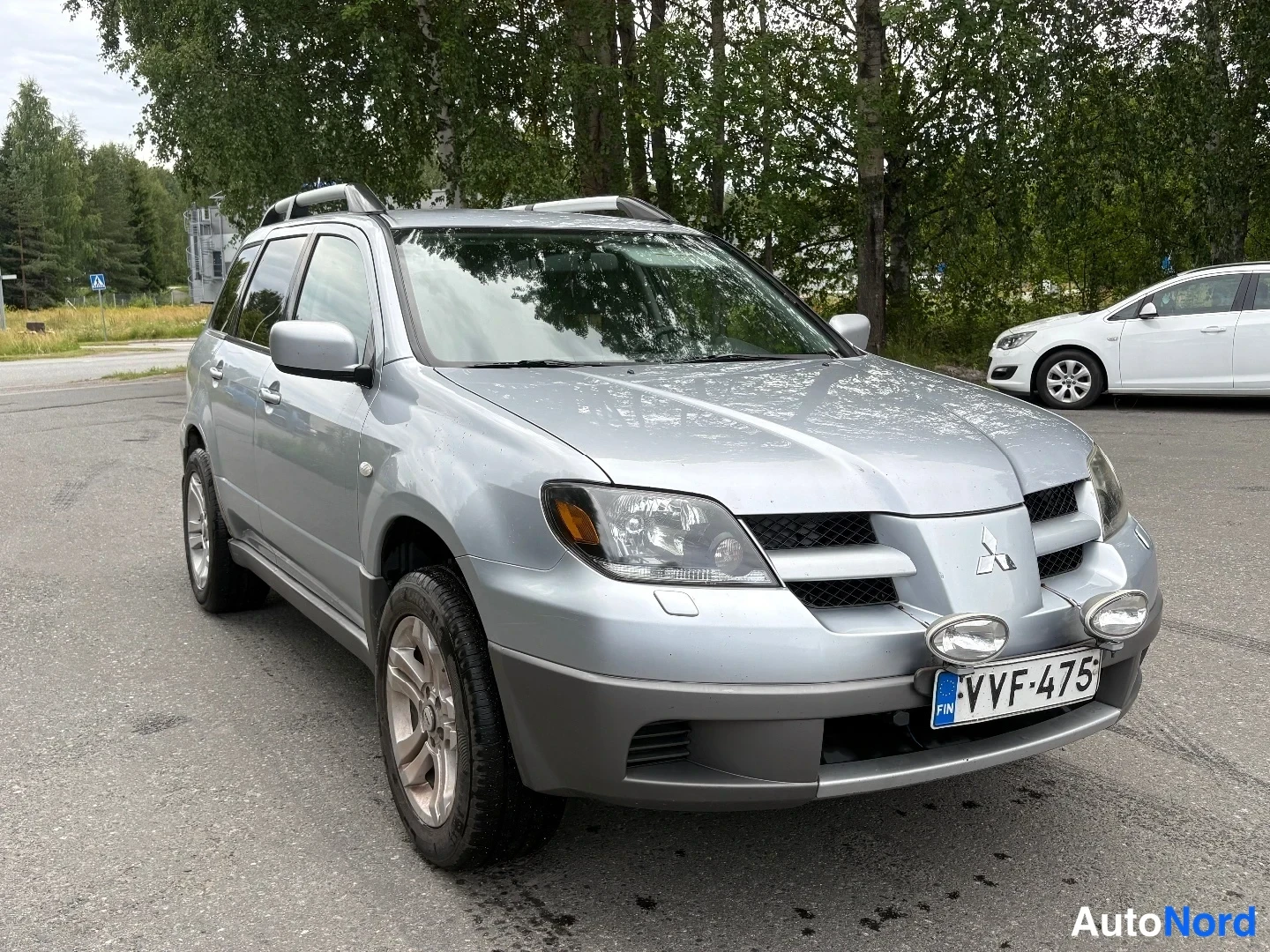 Etuviisto, nokka oikealle: Hopeanvärinen Mitsubishi Outlander kulmassa edestä oikealta, rekisteritunnus, kattokaiteet ja lisävalot puskurissa.