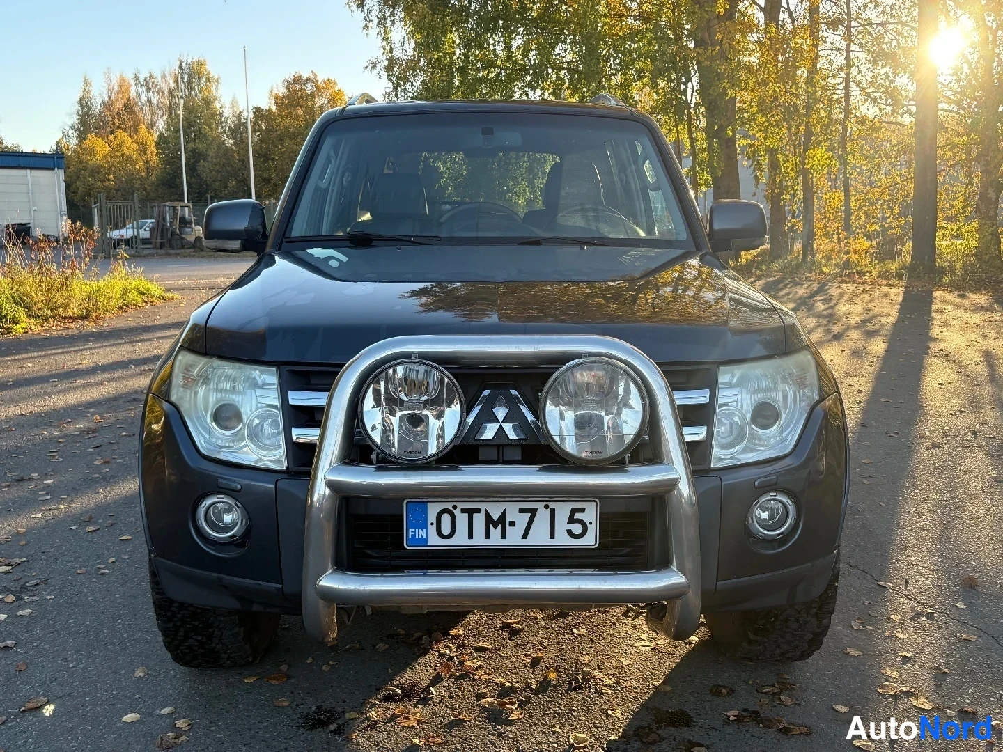 Edestä: Tummanharmaa Mitsubishi Pajero suoraan edestä kuvattuna, keulapuomi ja lisävalot, Mitsubishi-merkki ja rekisterikilpi.