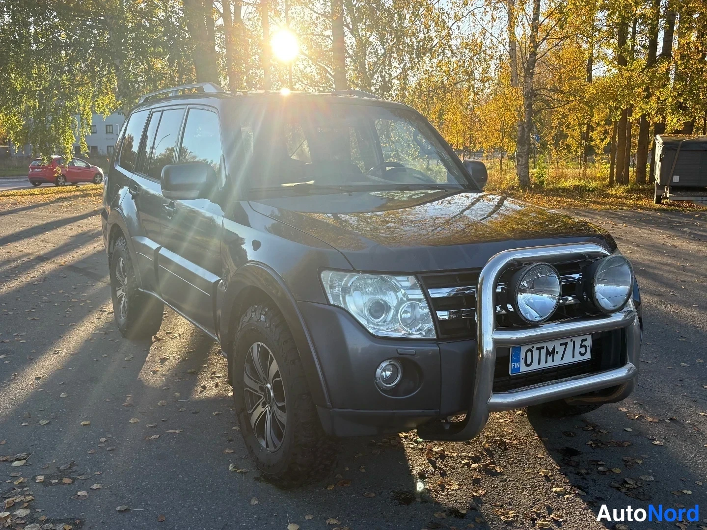 Etuviisto, nokka oikealle: Tummanharmaa Mitsubishi Pajero -maastoauto kolmannelta etukulmalta, keulapuomi ja lisävalot, rekisterikilpi, aurinko taustalla.