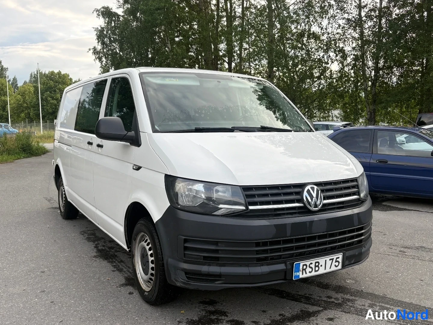 Etuviisto, nokka oikealle: Valkoinen Volkswagen Transporter -pakettiauto etukulmasta oikealta, musta etupuskuri, rekisterikilpi, takana sivuväylä.