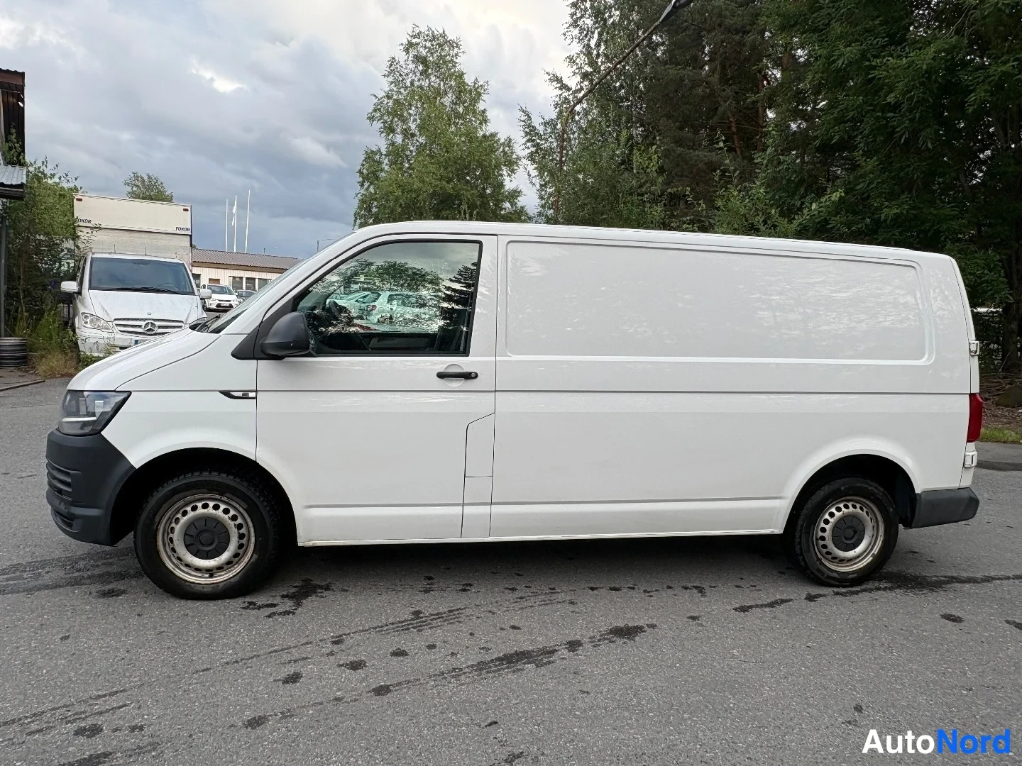 Sivulta: Valkoisen Volkswagen Transporter -pakettiauton sivunäkymä oikealta, umpipakettiauton kylki ilman takasivuikkunoita.