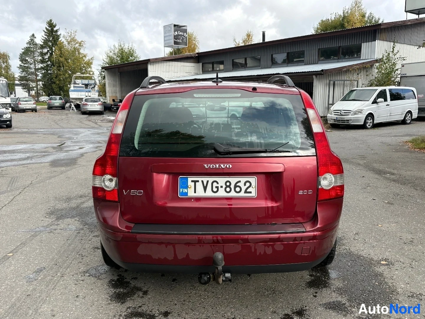 Takaa: Tummanpunaisen Volvo V50 -farmarin takaosa suoraan takaa; VOLVO- ja V50-merkinnät, rekisteritunnus ja vetokoukku näkyvissä.