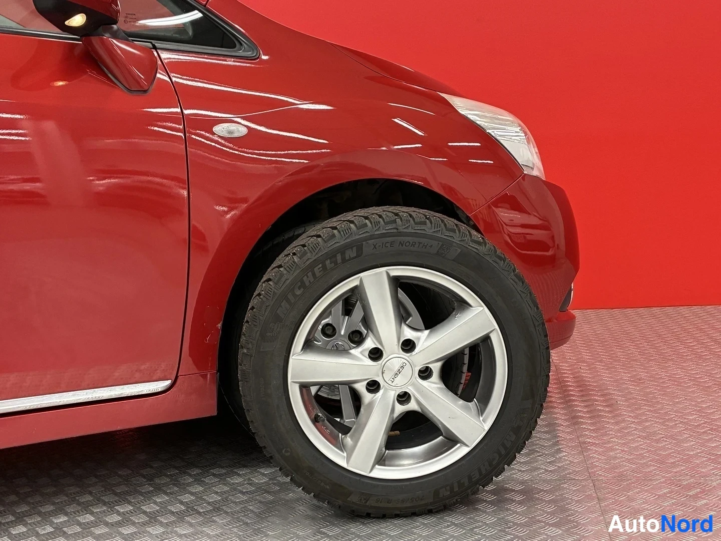 Etuviisto, nokka oikealle: Punaisen auton etukulma: etupyörä Michelin X-Ice North -renkaalla ja hopeinen vanteen pölykki punaisen seinän edessä.