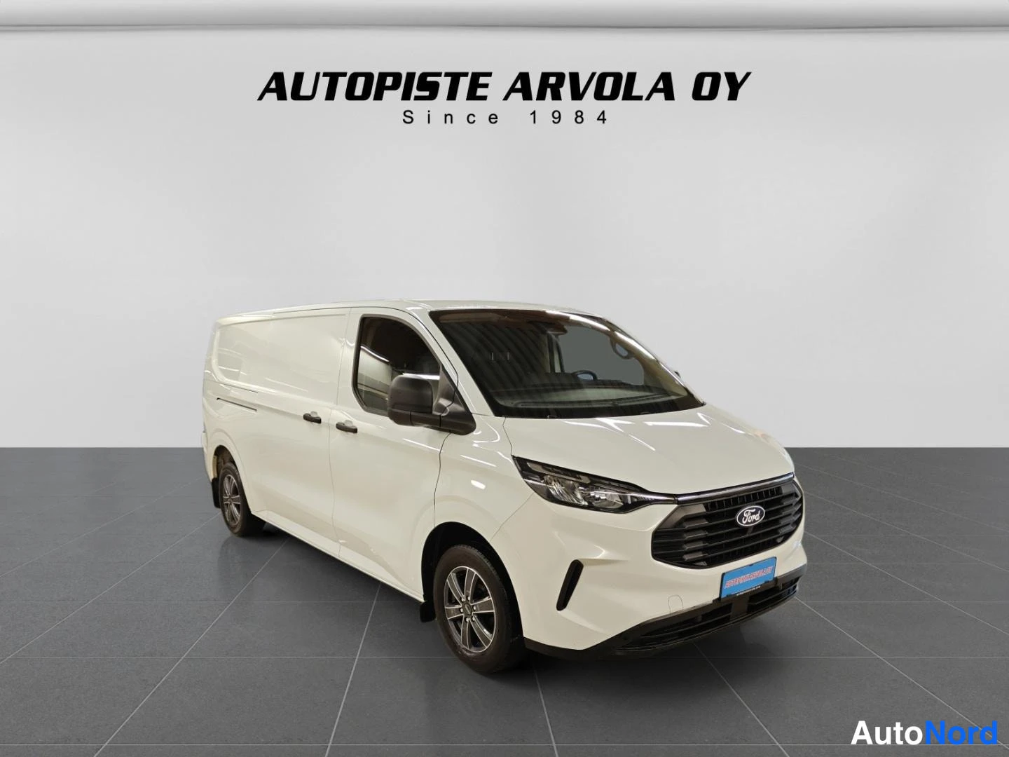 Etuviisto, nokka oikealle: Ford Transit Custom -pakettiauto etuviistosta oikealta, valkoinen koripinta näkyy kokonaan studiotaustaa vasten.