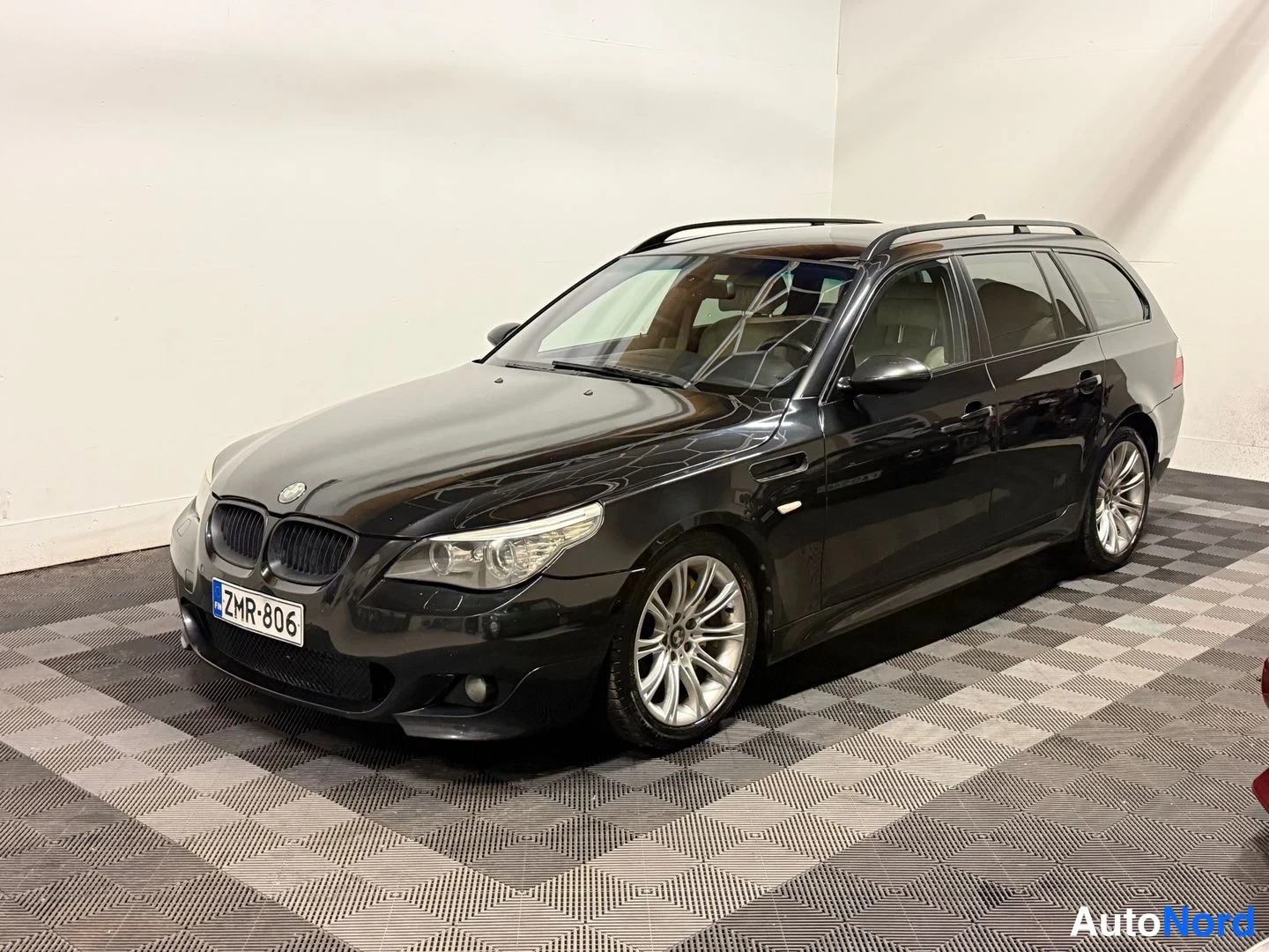 Etuviisto, nokka vasemmalle: Mustan Farmarin BMW:n etukulmanäkymä sisätiloissa; näkyvissä etupanssari, kattokaiteet ja -rekisterikilpi.