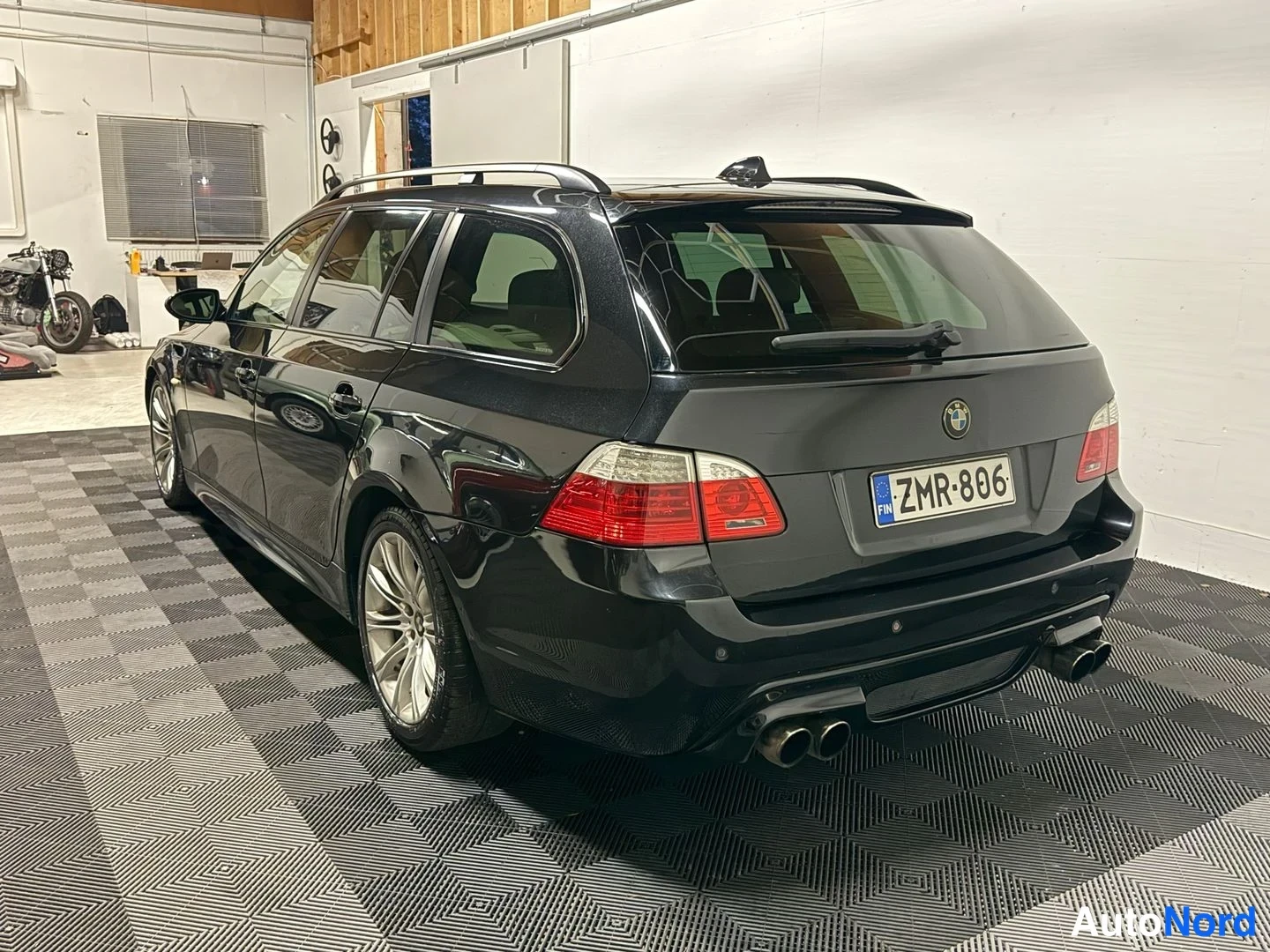 Takaviisto: Mustan BMW:n takakulmanäkymä sisätiloissa; näkyvissä takavalojen ja rekisterikilven lisäksi neljä pakoputkea ja kattokaiteet.