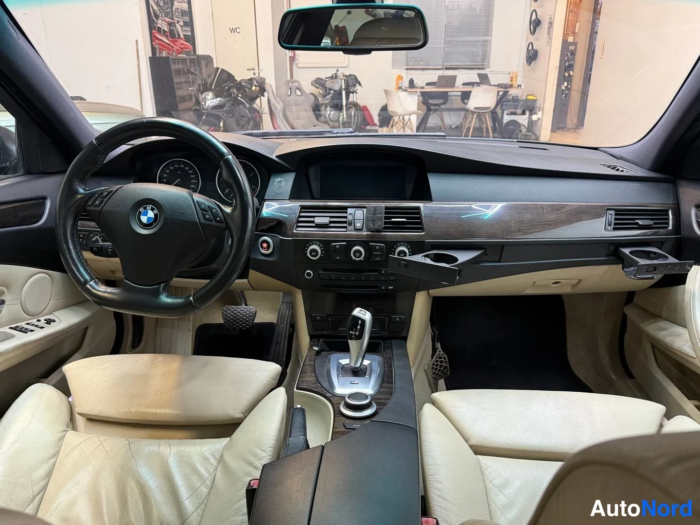 Sivulta: BMW:n ohjaamo nähtynä takaistuimelta; näkyvissä BMW-ohjauspyörä, tumma kojelauta, puukoristeet ja vaaleat nahkaistuimet.