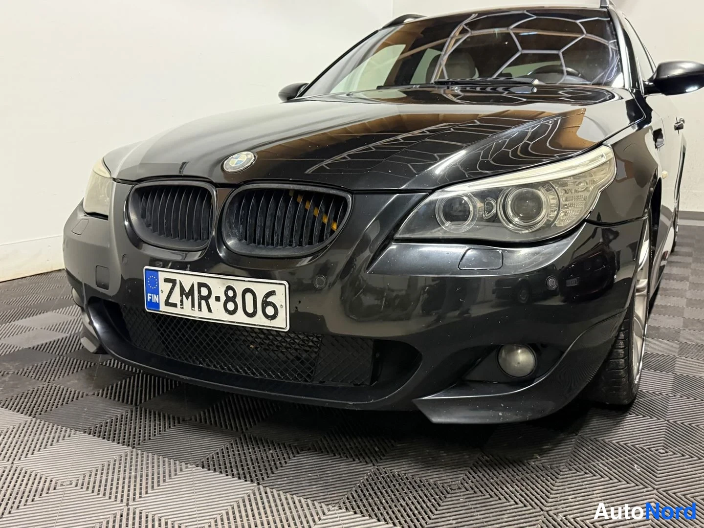 Etuviisto, nokka vasemmalle: Mustan BMW:n etukolmiveto: etusäleikkö, ajovalot ja rekisterikilpi kuvattuna sisätiloissa.