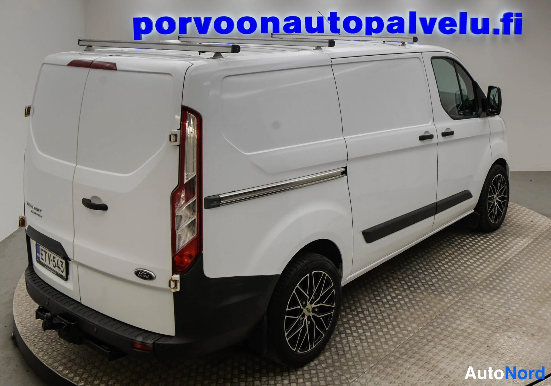 Takaviisto: Valkoinen Ford Transit Custom -pakettiauto kolmasta neljänneksestä takaa vasemmalti katsottuna, kattotelineet ja sivuportaat näkyvissä.