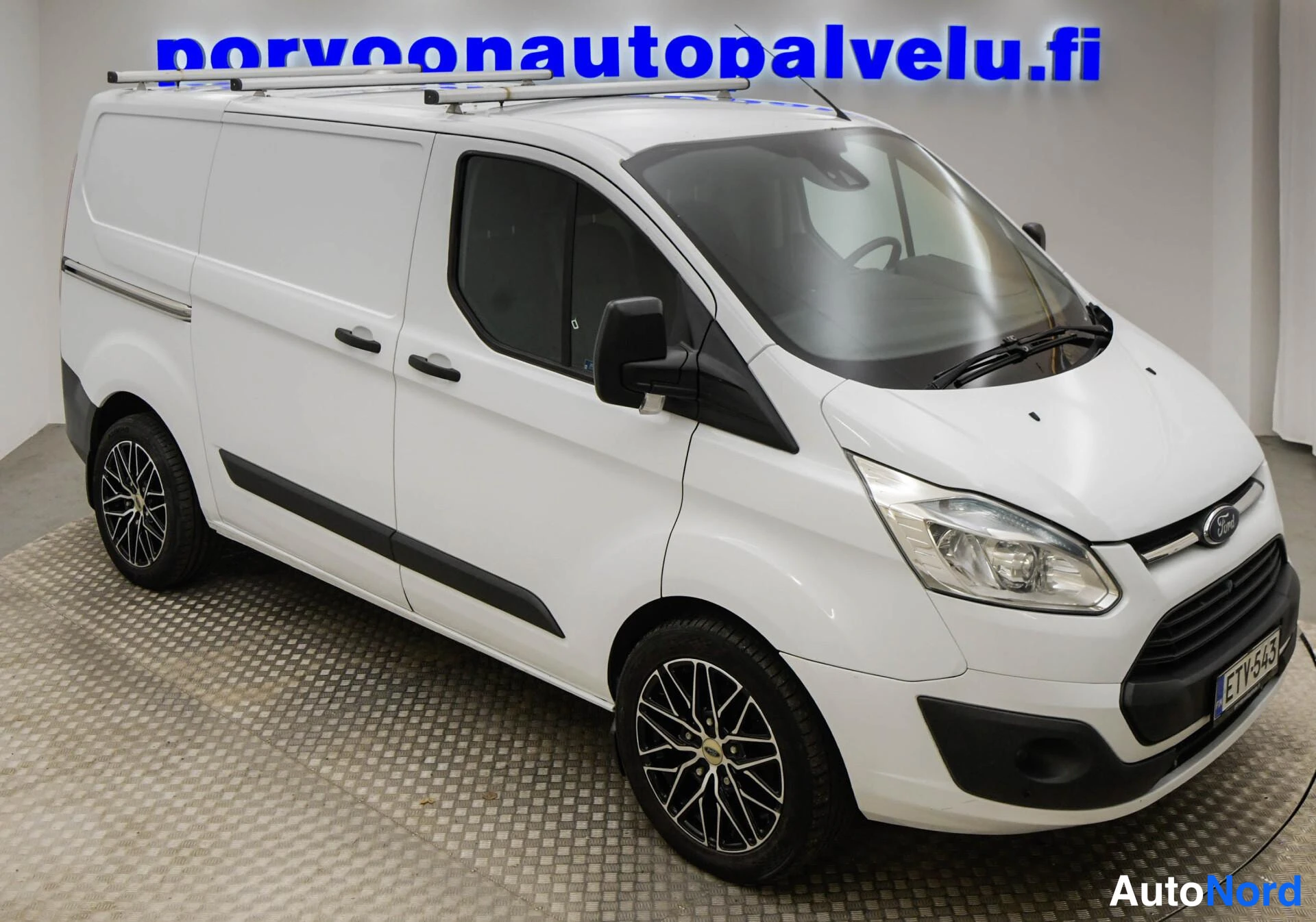 Etuviisto, nokka vasemmalle: Valkoinen Ford Transit Custom etuviistossa; etugrilli ja Ford-merkki näkyvät vasemmalla ja takapyörä oikealla. Rekisterikilpi, kattotelineet, tummat va