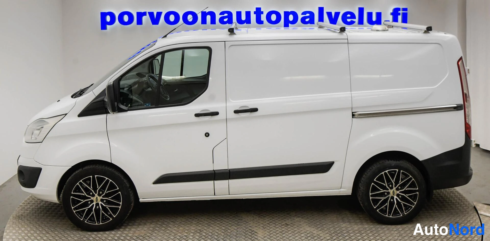 Etuviisto, nokka vasemmalle: Valkoisen Ford Transit Customin sivukulma peilisuunnassa; grillin reuna vasemmalla ja takapyörä oikealla osoittavat nokan olevan vasemmalla. Kattotelin